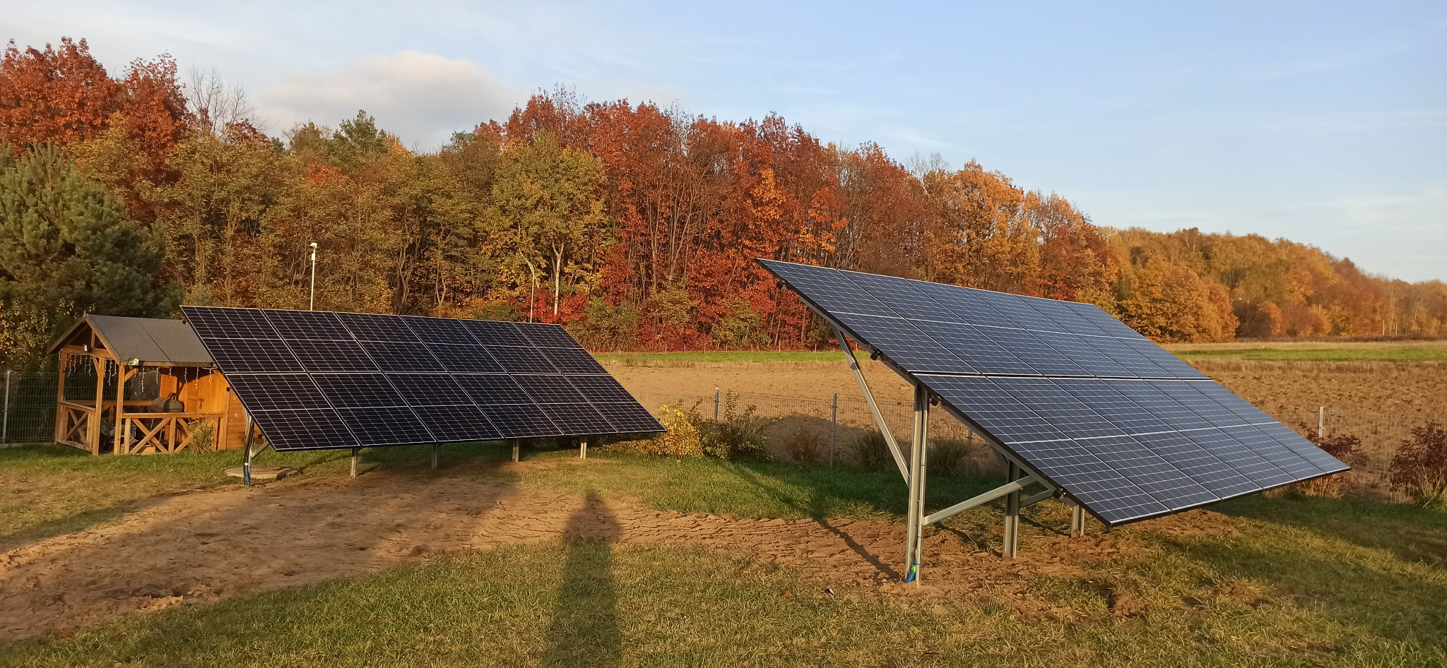 Dwa panele fotowoltaiczne na tle jesiennego lasu i pola, cień osoby w dolnej części kadru.