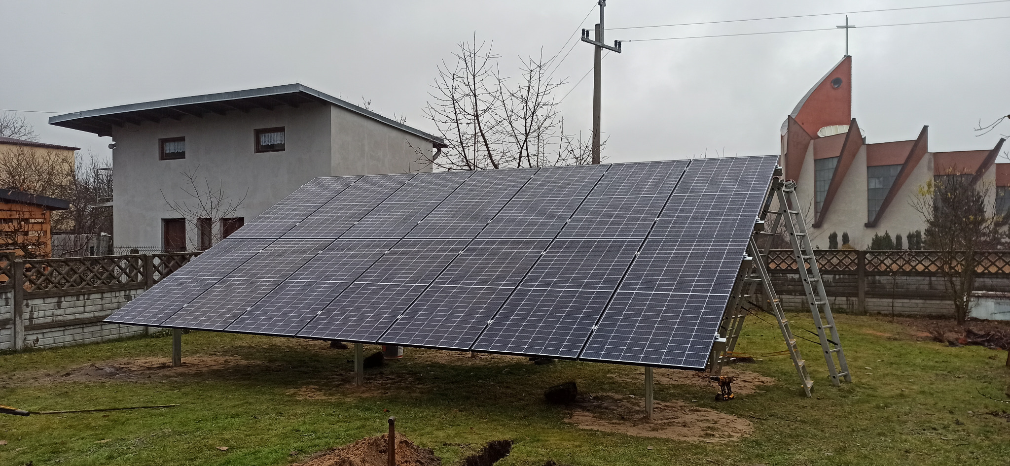 Montaż paneli słonecznych na konstrukcji wsporczej w ogrodzie, w tle dom i kościół, widoczna drabina i narzędzia.