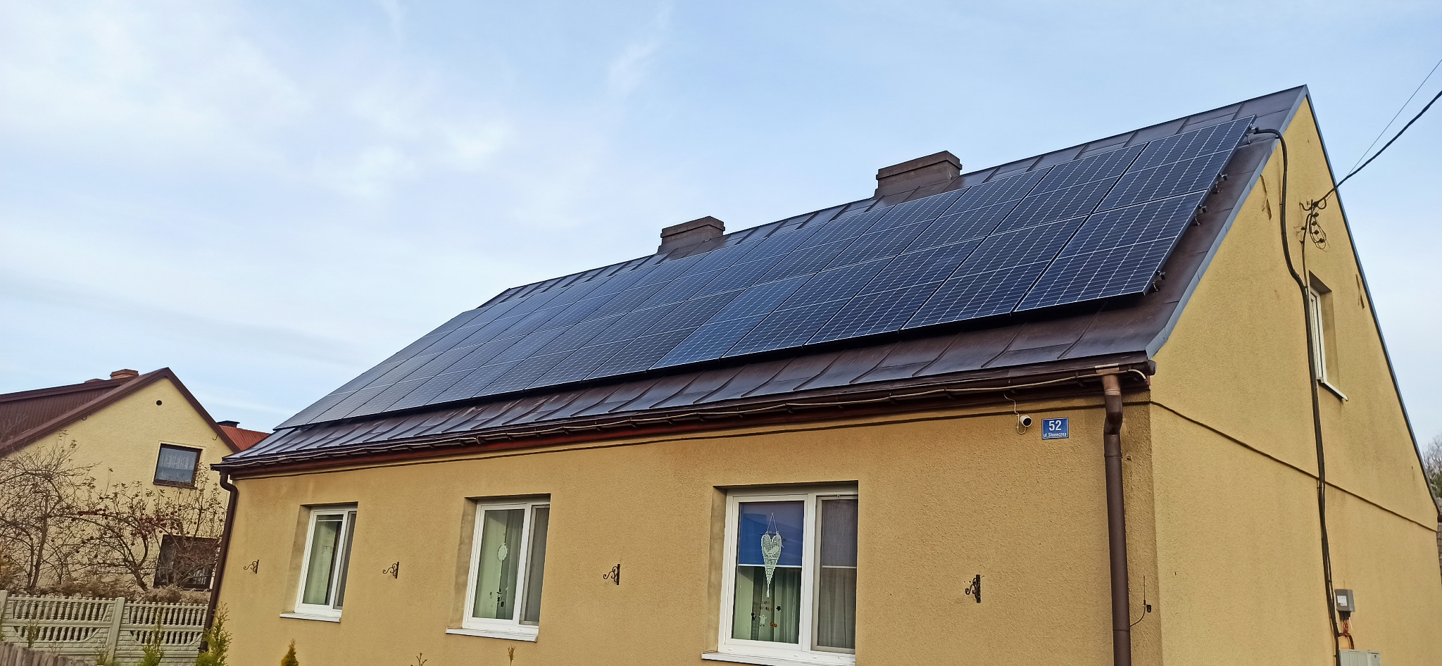 Dom jednorodzinny z panelami słonecznymi na brązowym dachu, widoczny numer domu '52' i ozdobne elementy pod oknami.