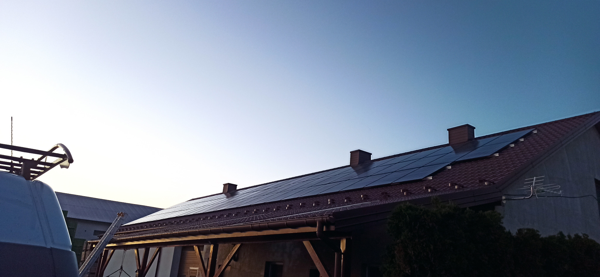 Panele słoneczne na dachu domu z brązową dachówką, widoczne kominy i antena telewizyjna, na pierwszym planie fragment samochodu dostawczego z bagażnikiem dachowym i drabina.