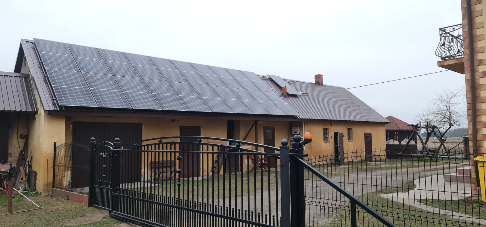 Instalacja paneli słonecznych na dachu budynku gospodarczego z żółtą elewacją, widok zza czarnego, metalowego ogrodzenia.