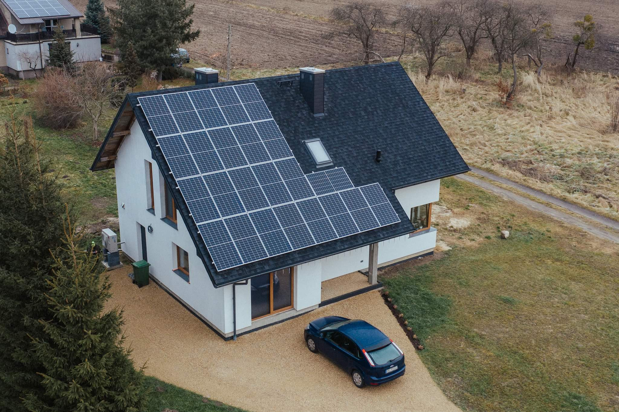 Widok z góry na biały dom jednorodzinny z ciemnym dachem pokrytym panelami słonecznymi; na podjeździe stoi niebieski samochód osobowy.