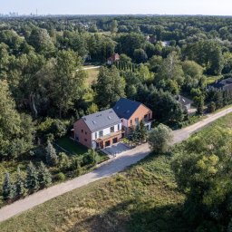EVEN SP. z o.o. - Widok z lotu ptaka na nowoczesny, dwukondygnacyjny dom z płaskim dachem, wykończony cegłą i białym tynkiem, otoczony zielenią, z widoczną panoramą miasta w oddali.