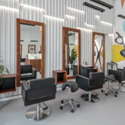 Wnętrze nowoczesnego salonu fryzjerskiego z czterema stanowiskami: czarne fotele na srebrnych podstawach, drewniane ramy luster, białe ściany z podświetleniem LED i mural z motywem oka i nożyczek.