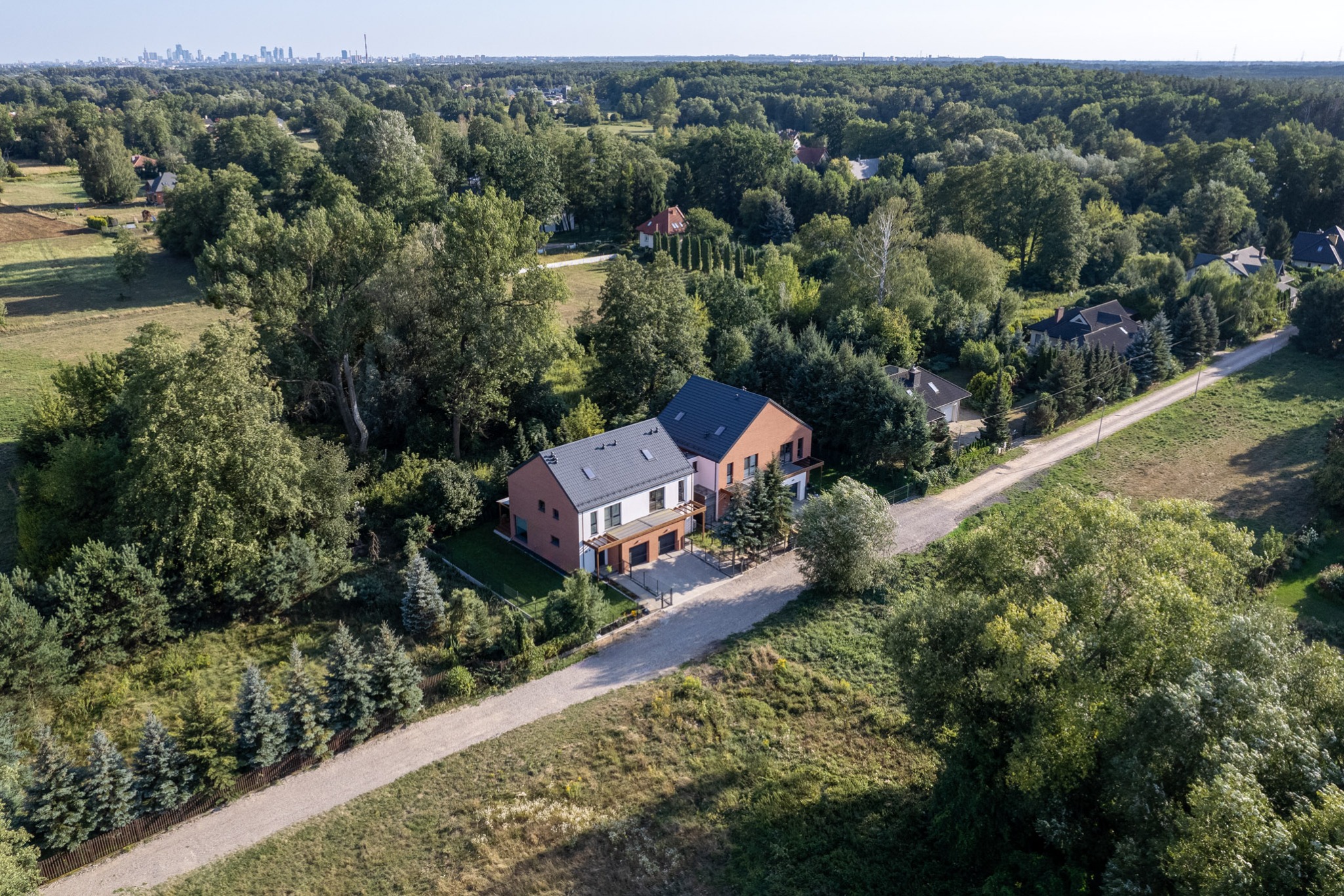 Widok z lotu ptaka na nowoczesny, dwukondygnacyjny dom z płaskim dachem, wykończony cegłą i białym tynkiem, otoczony zielenią, z widoczną panoramą miasta w oddali.