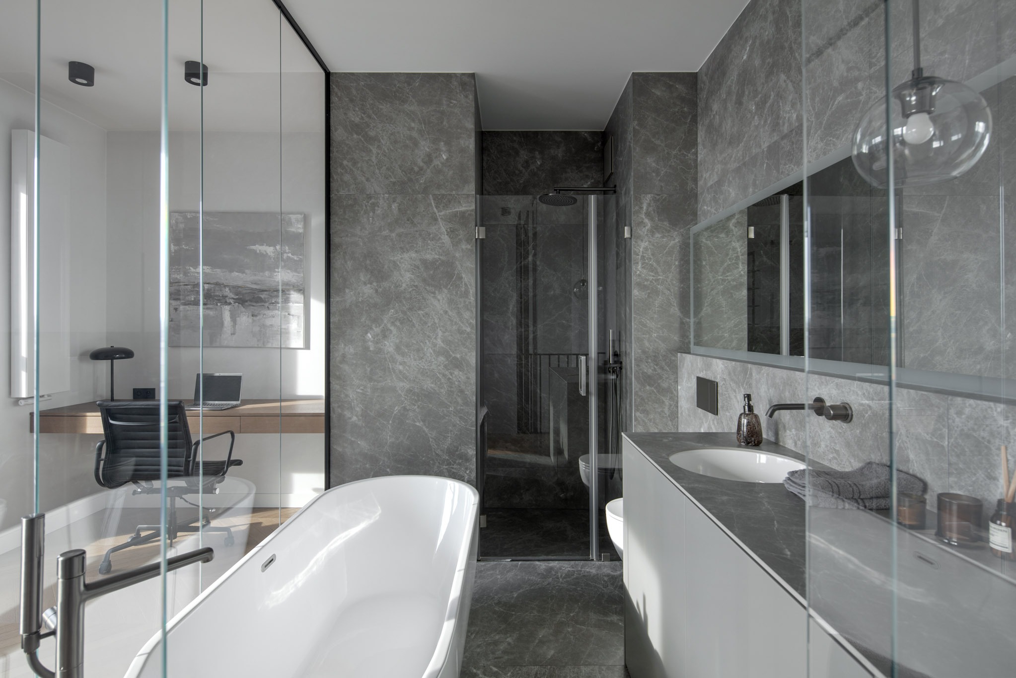 Nowoczesna łazienka z wanną wolnostojącą, kabiną prysznicową i biurkiem w tle, oddzielona szklaną ścianą. Szare płytki imitujące marmur na ścianach i podłodze.