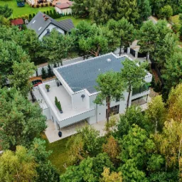 Nowoczesny, biały dom z płaskim dachem otoczony zielenią, widok z lotu ptaka, z tarasem na dachu, gdzie stoją donice z roślinami.