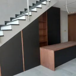 Nowoczesna zabudowa pod schodami z czarnymi frontami i drewnianymi akcentami, w tle widoczna szafa i drzwi w podobnym stylu, szare ściany.
