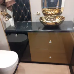 Nowoczesna łazienka z czarnym blatem z połyskiem, złotą umywalką nablatową, szafką z połyskującymi frontami i kryształowymi uchwytami oraz toaletą w zabudowie.