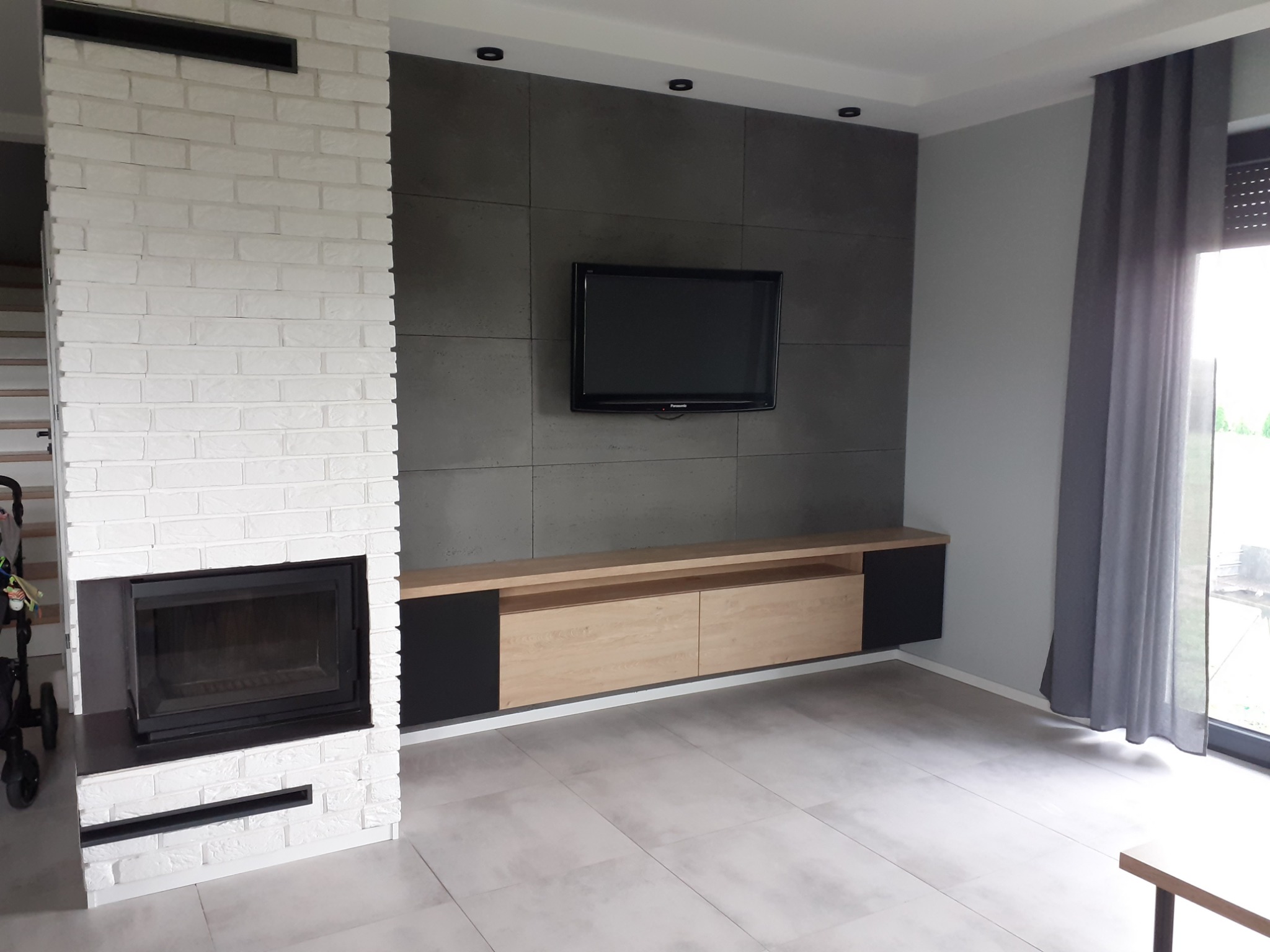 Nowoczesny salon z kominkiem obudowanym białą cegłą, szarą ścianą z telewizorem i szafką RTV z jasnego drewna z czarnymi elementami.