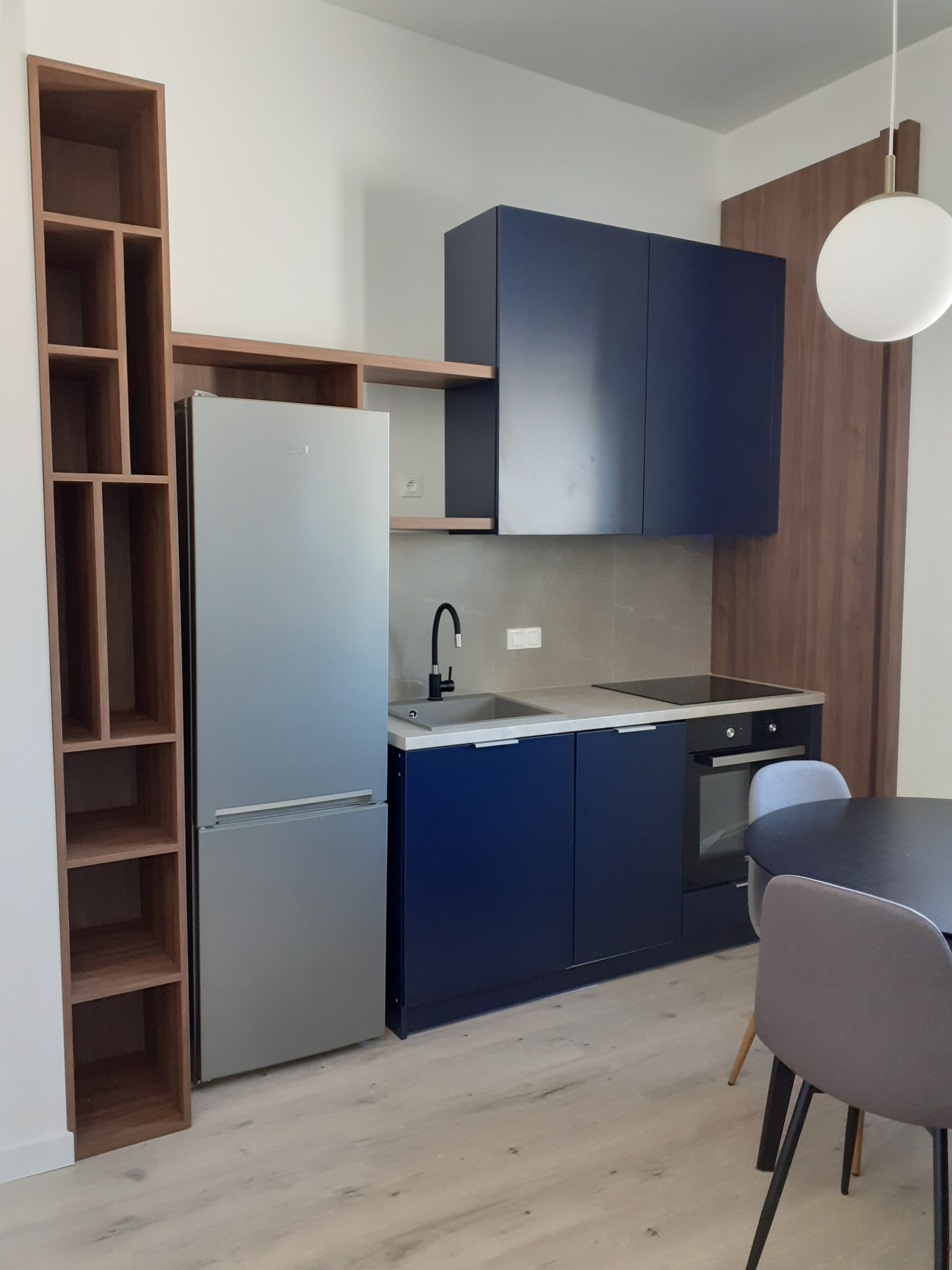 Nowoczesna kuchnia z granatowymi szafkami, szarą ścianką nad blatem, srebrną lodówką i drewnianym regałem z otwartymi półkami, uzupełniona okrągłym stołem z szarymi krzesłami.