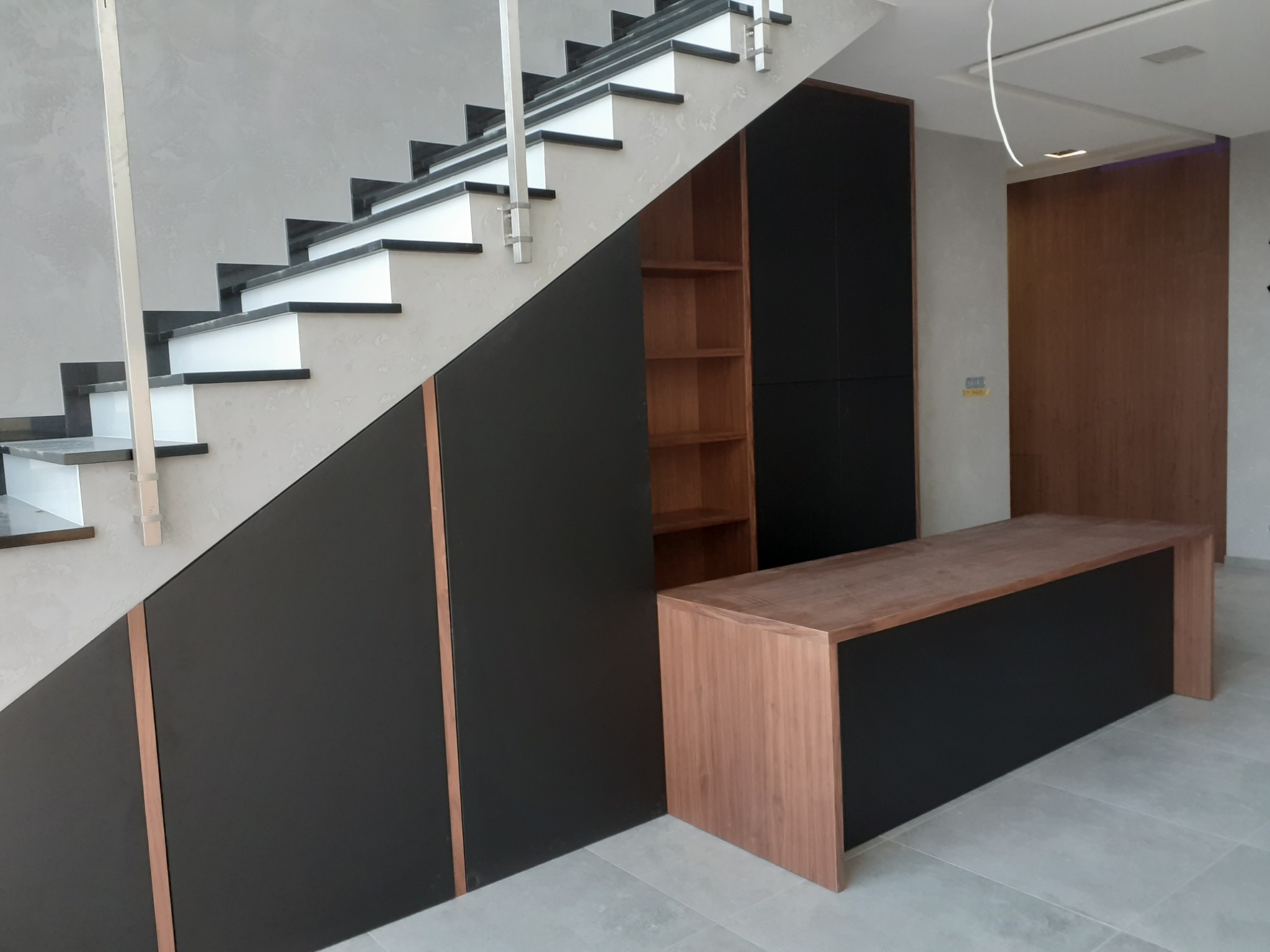 Nowoczesna zabudowa pod schodami z czarnymi frontami i drewnianymi akcentami, w tle widoczna szafa i drzwi w podobnym stylu, szare ściany.