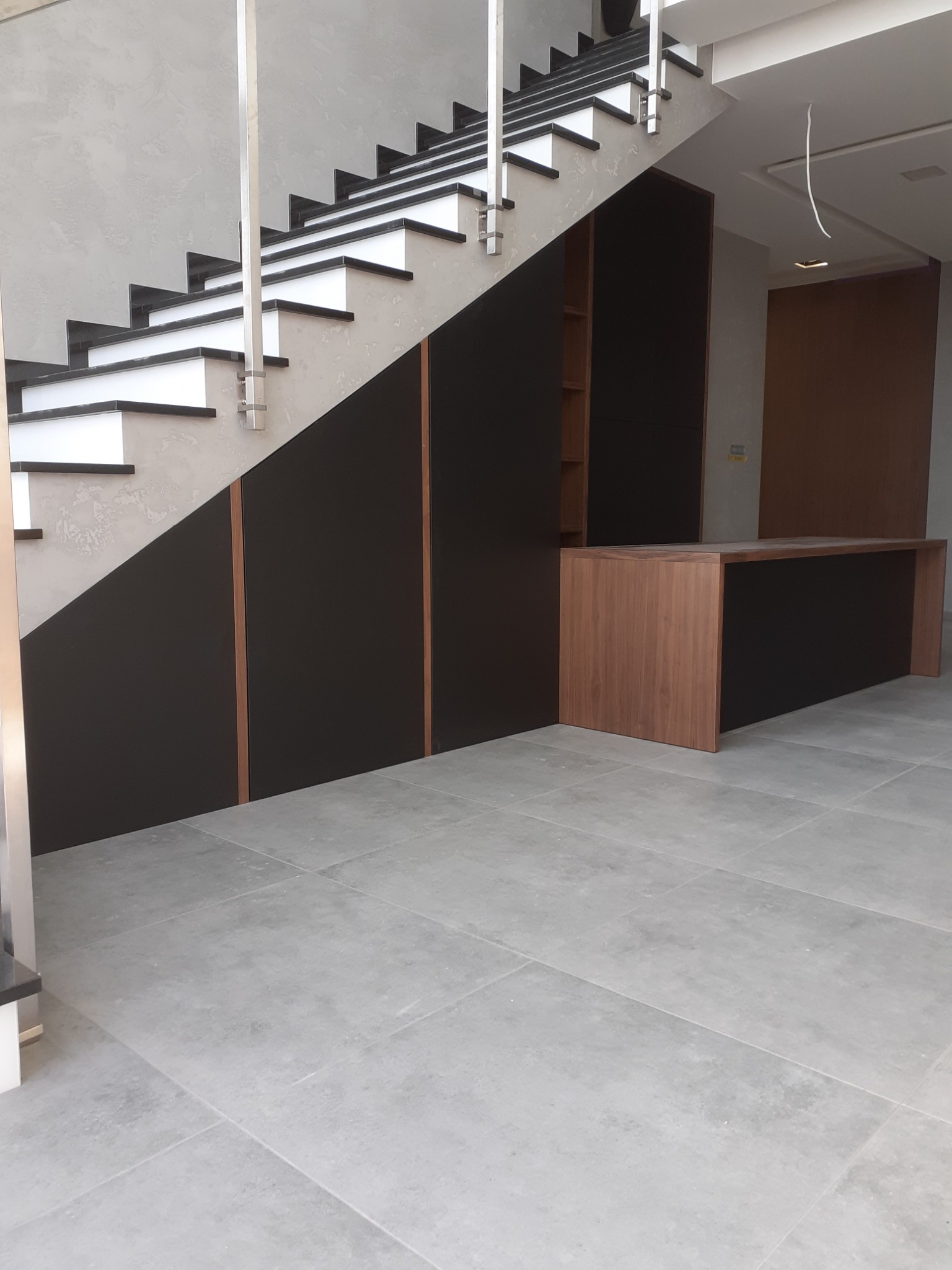 Nowoczesne schody z zabudową szaf wnękowych pod nimi oraz drewnianą konsolą. Minimalistyczny design, szara podłoga i ściany.