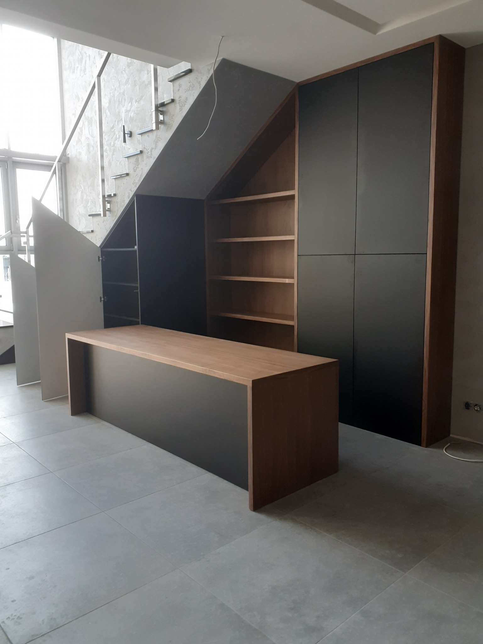 Zabudowa pod schodami: szafa z czarnymi frontami, regał z drewnianymi półkami i blat z drewna na czarnej podstawie, widok na surowy beton na ścianie i schody.