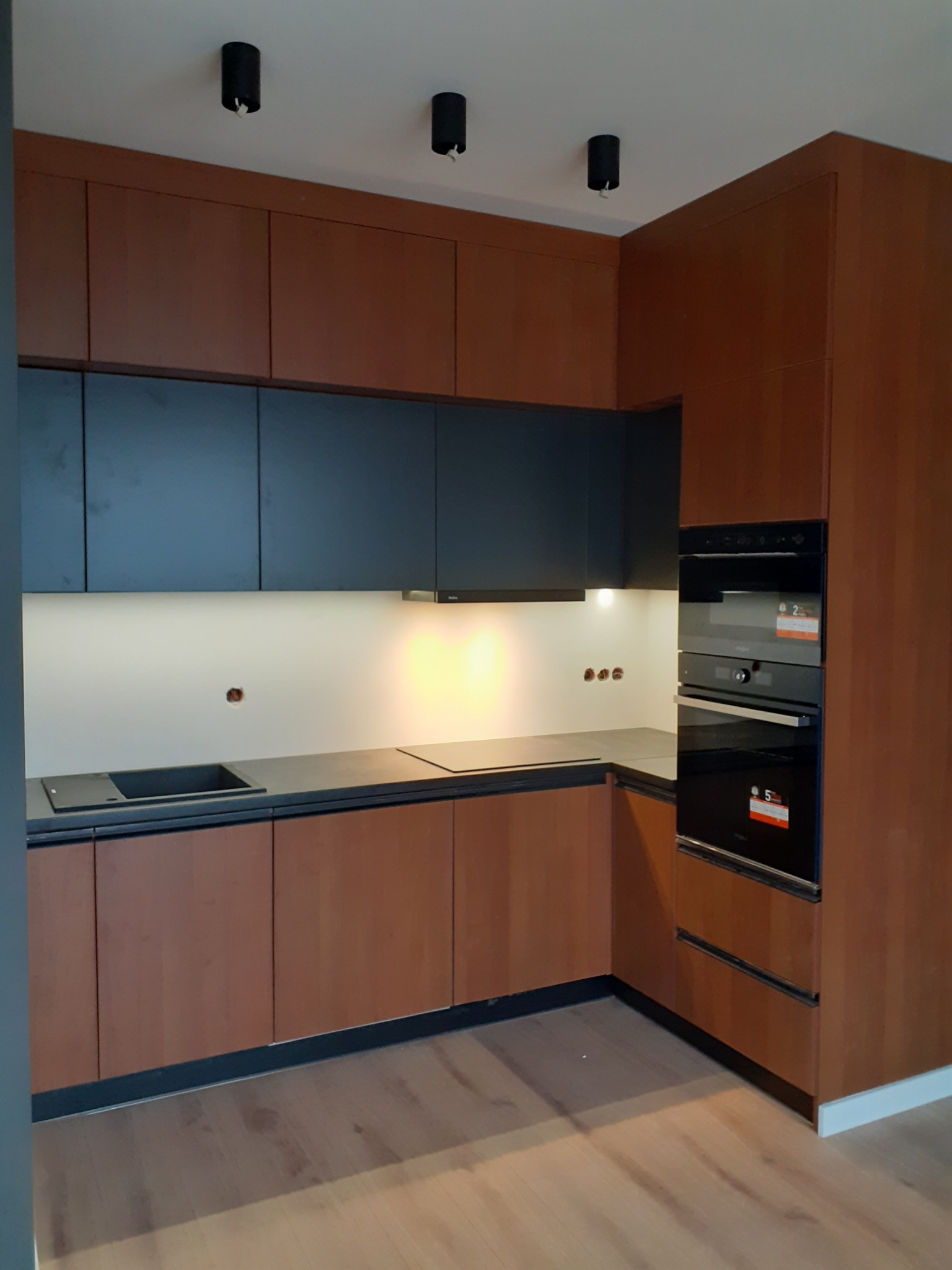 Nowoczesna kuchnia z drewnianymi frontami szafek dolnych i czarnymi szafkami górnymi, czarny blat, zabudowane piekarniki i minimalistyczne oświetlenie sufitowe.