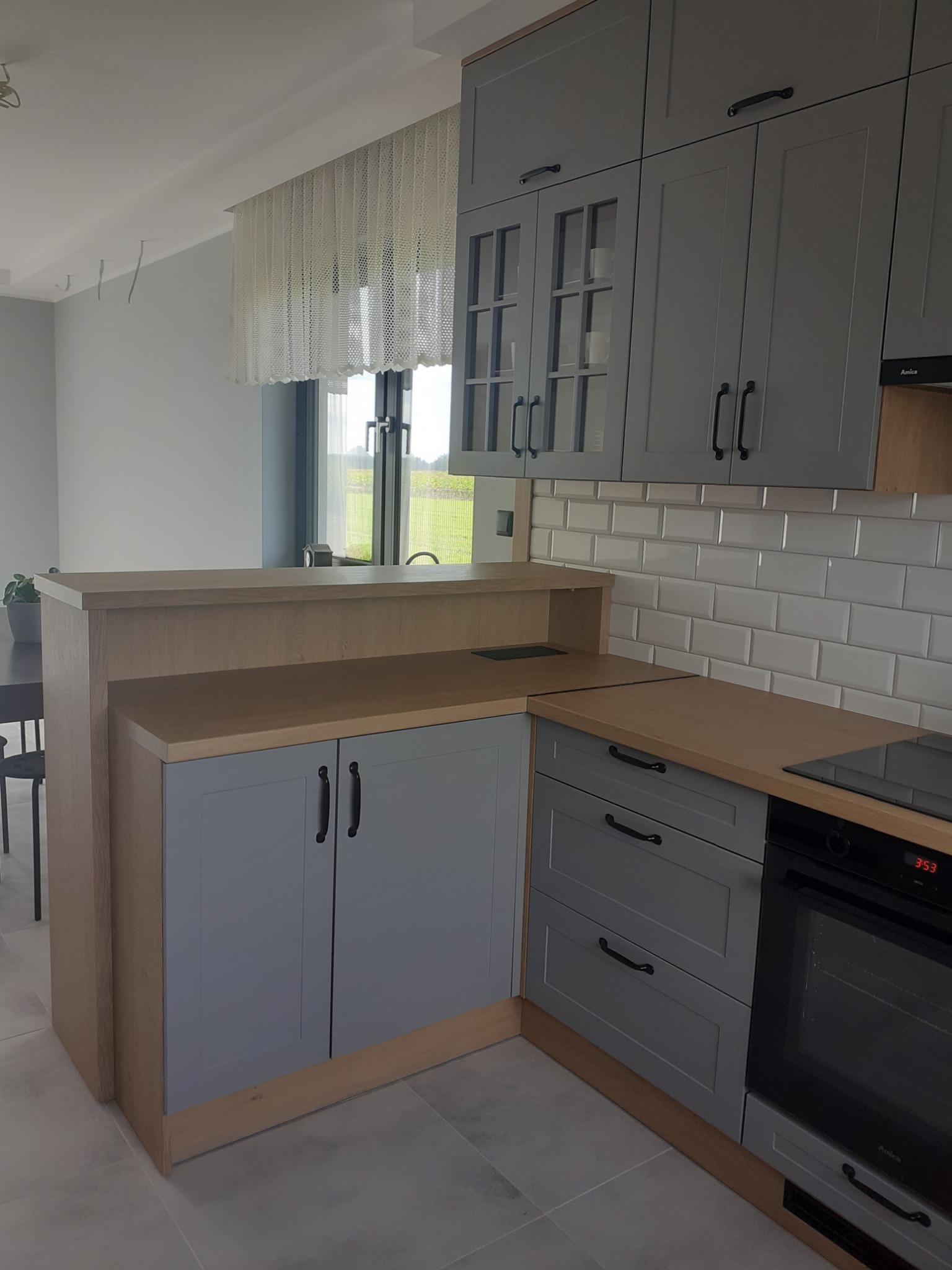 Szare szafki kuchenne z drewnianym blatem, fragment zabudowy z piekarnikiem i barkiem, białe kafelki na ścianie.