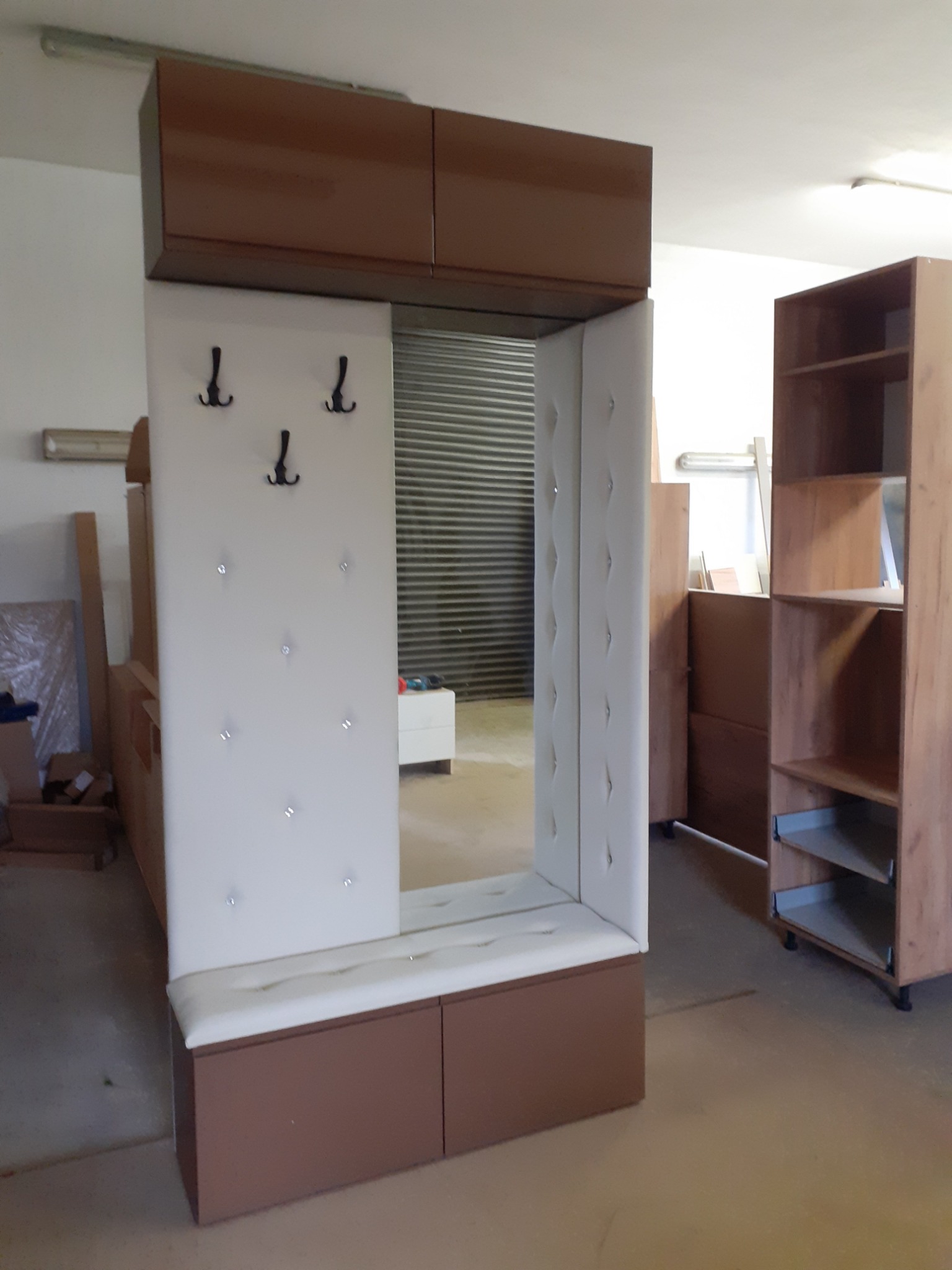 Komplet mebli do przedpokoju: szafka z siedziskiem, lustro z wieszakami i górna szafka w kolorze brązowo-białym, widoczny regał z półkami w tle.