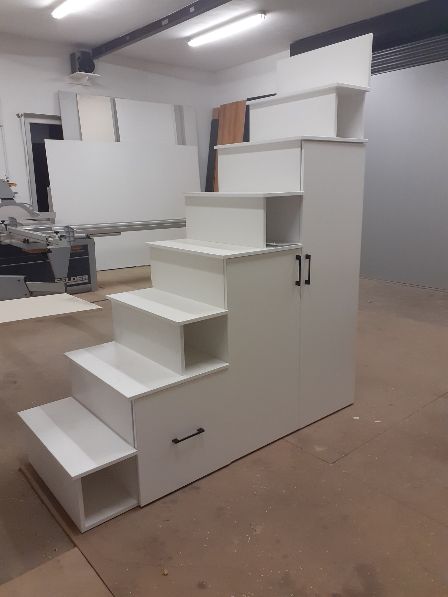 Biała konstrukcja schodkowa z szafkami i szufladą, prawdopodobnie do pokoju dziecięcego, widoczna w warsztacie stolarskim, obok maszyna do obróbki drewna.