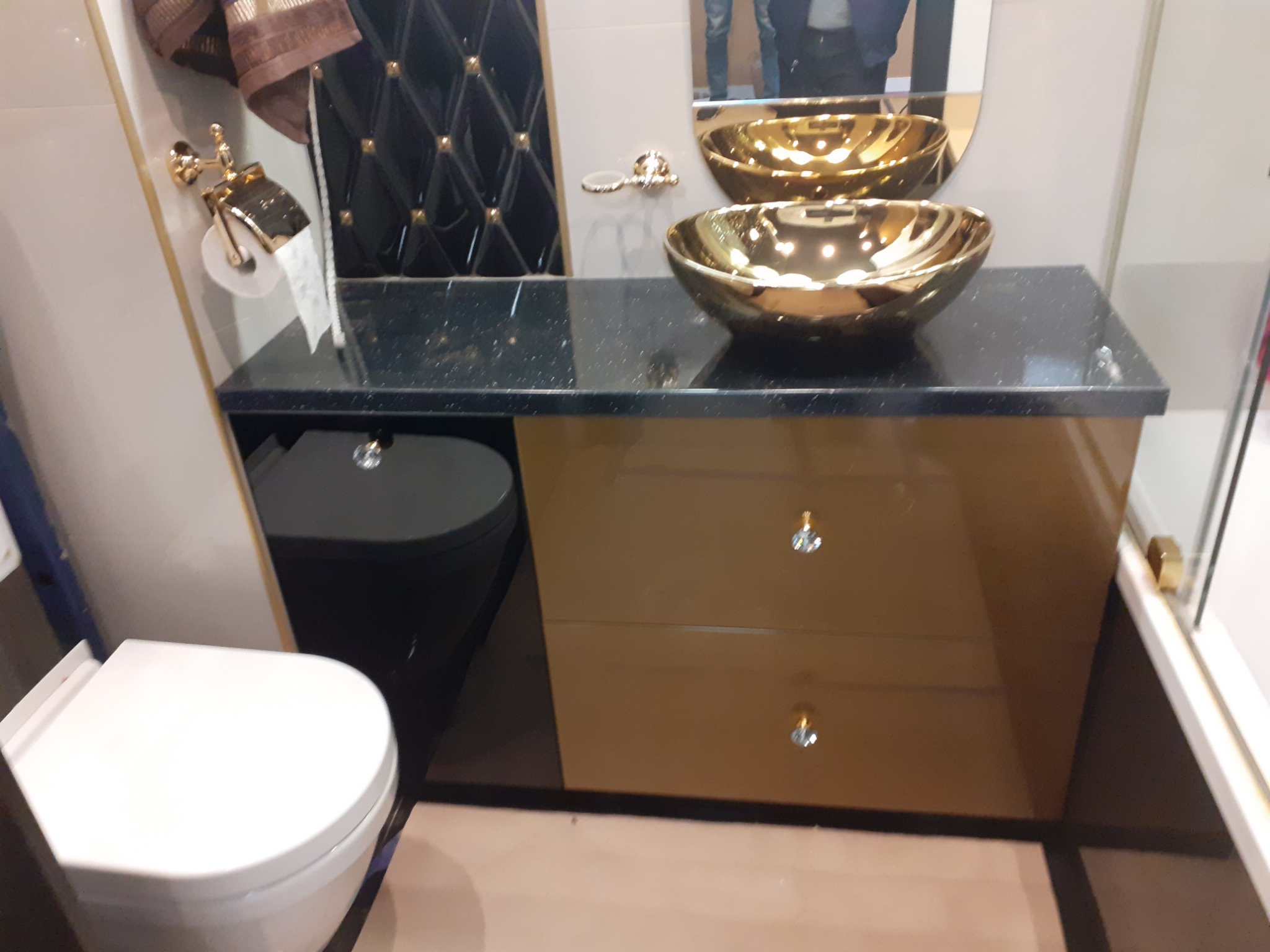 Nowoczesna łazienka z czarnym blatem z połyskiem, złotą umywalką nablatową, szafką z połyskującymi frontami i kryształowymi uchwytami oraz toaletą w zabudowie.