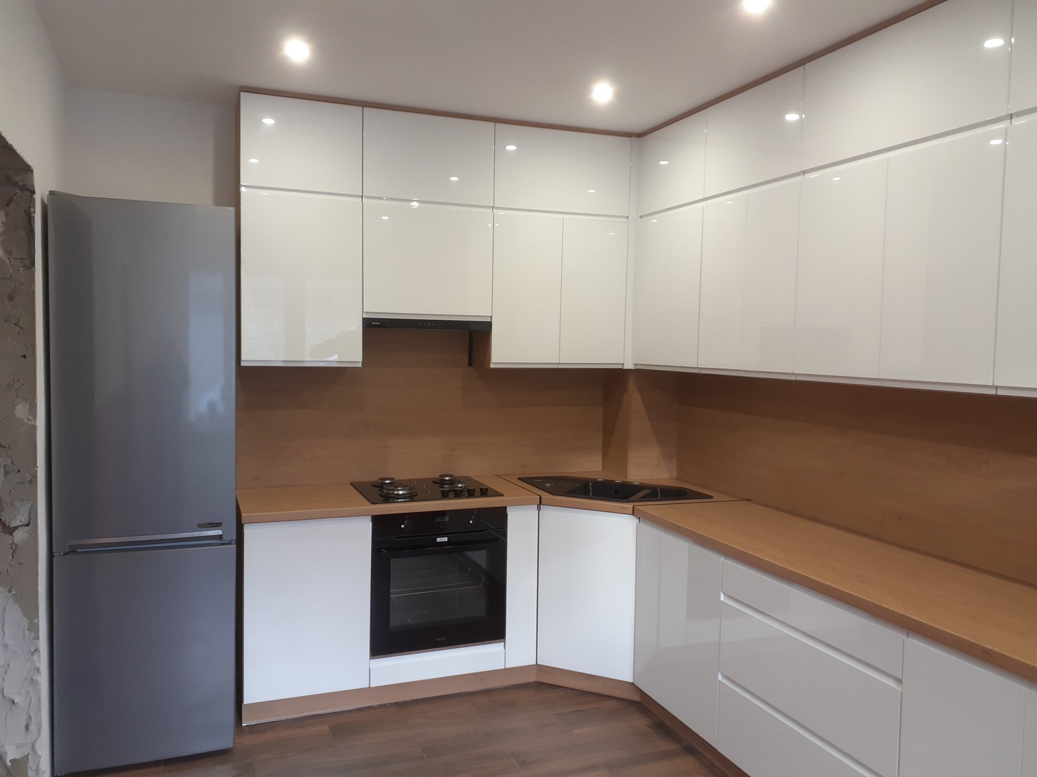 Nowoczesna kuchnia z białymi, lakierowanymi frontami, drewnianym blatem i szarą lodówką wolnostojącą, widoczna ściana w trakcie wykończenia.