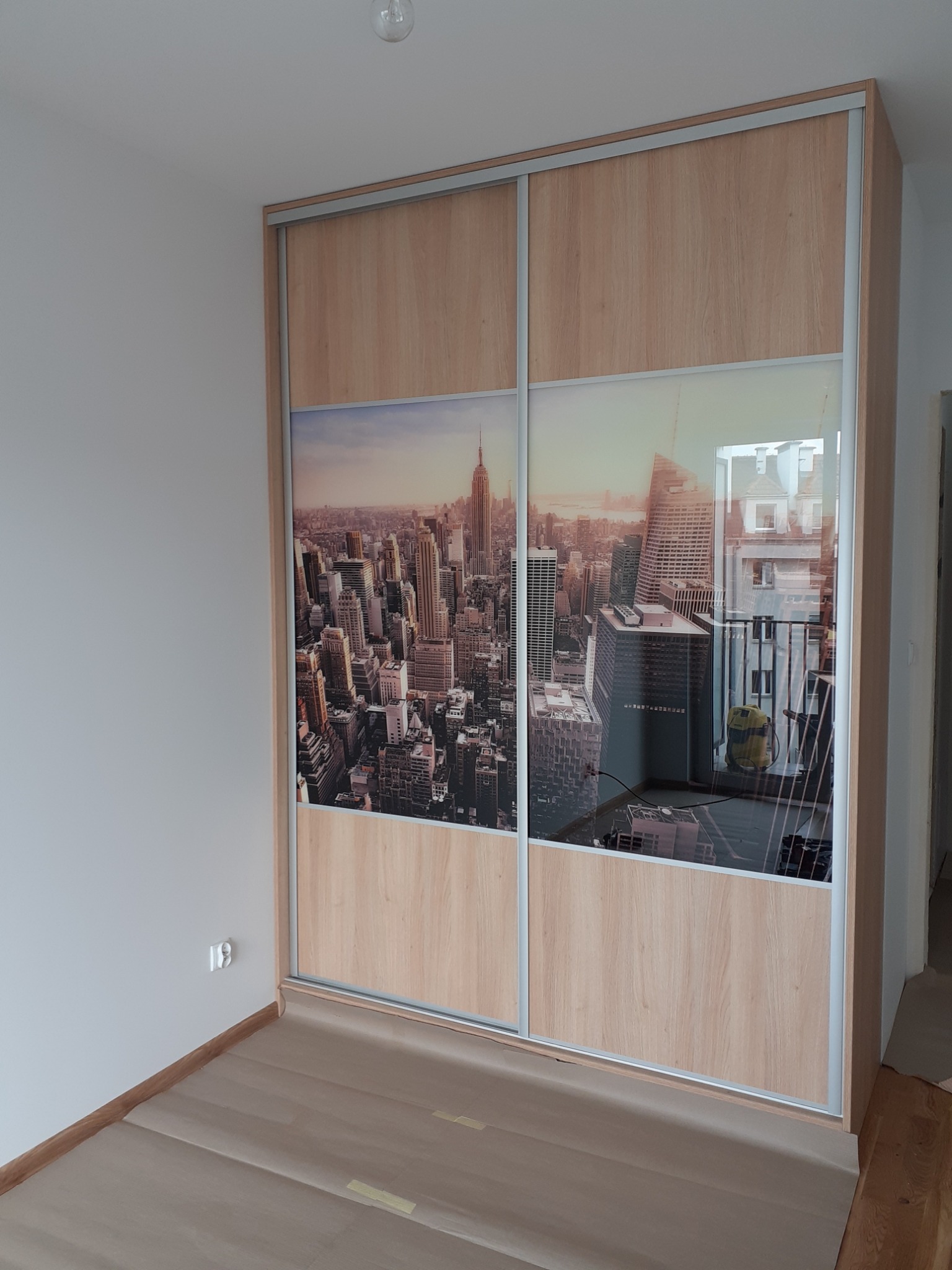 Szafa przesuwna z okładziną imitującą drewno, z fototapetą przedstawiającą panoramę Nowego Jorku, widoczna w rogu pokoju z zabezpieczoną podłogą.