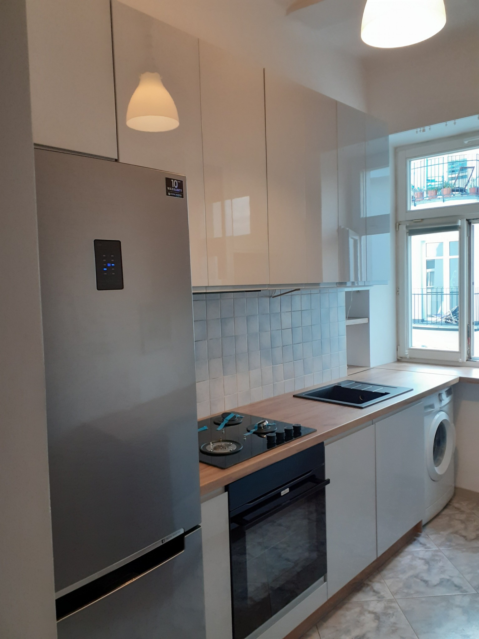 Nowoczesna kuchnia z białymi, błyszczącymi szafkami, drewnianym blatem, srebrną lodówką z wyświetlaczem, czarną płytą grzewczą i piekarnikiem oraz pralką pod oknem.