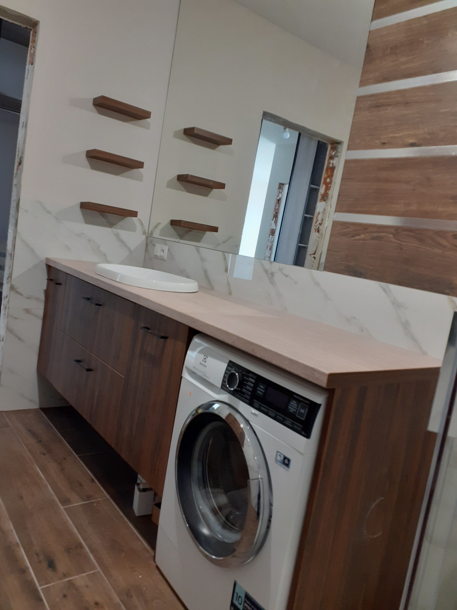 Łazienka z umywalką nablatową na szafce z ciemnego drewna, pralka zabudowana w szafce, lustro z półkami i marmurowe płytki na ścianie.