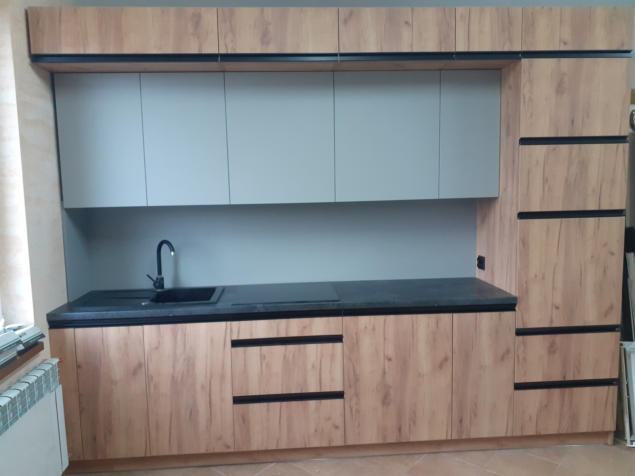 Nowoczesna zabudowa kuchenna z drewnopodobnymi frontami, szarymi szafkami wiszącymi i czarnym blatem oraz zlewem w kolorze czarnym.