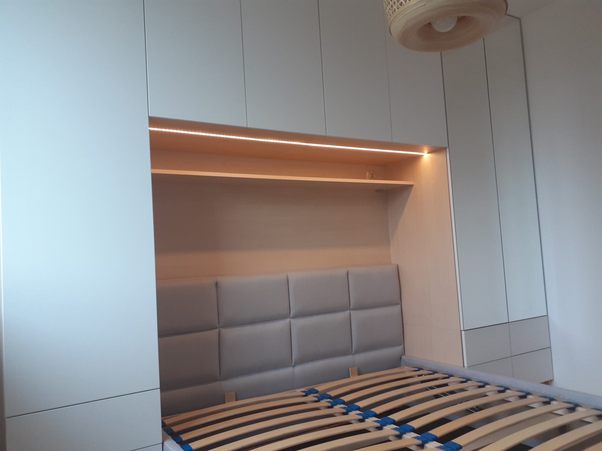 Szafa wnękowa z wbudowanym łóżkiem i podświetleniem LED, zagłówek z tapicerowanymi panelami w kolorze szarym, drewniany stelaż łóżka z niebieskimi elementami.