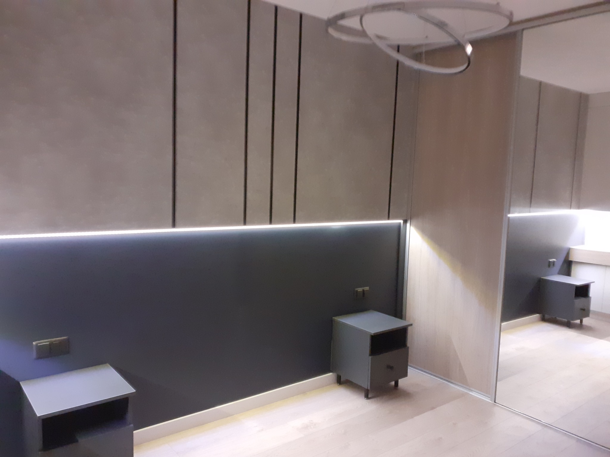 Nowoczesna sypialnia z szarą ścianą z pionowymi panelami, podświetleniem LED i szafkami nocnymi w kolorze szarym, odbicie w lustrzanych drzwiach szafy wnękowej.