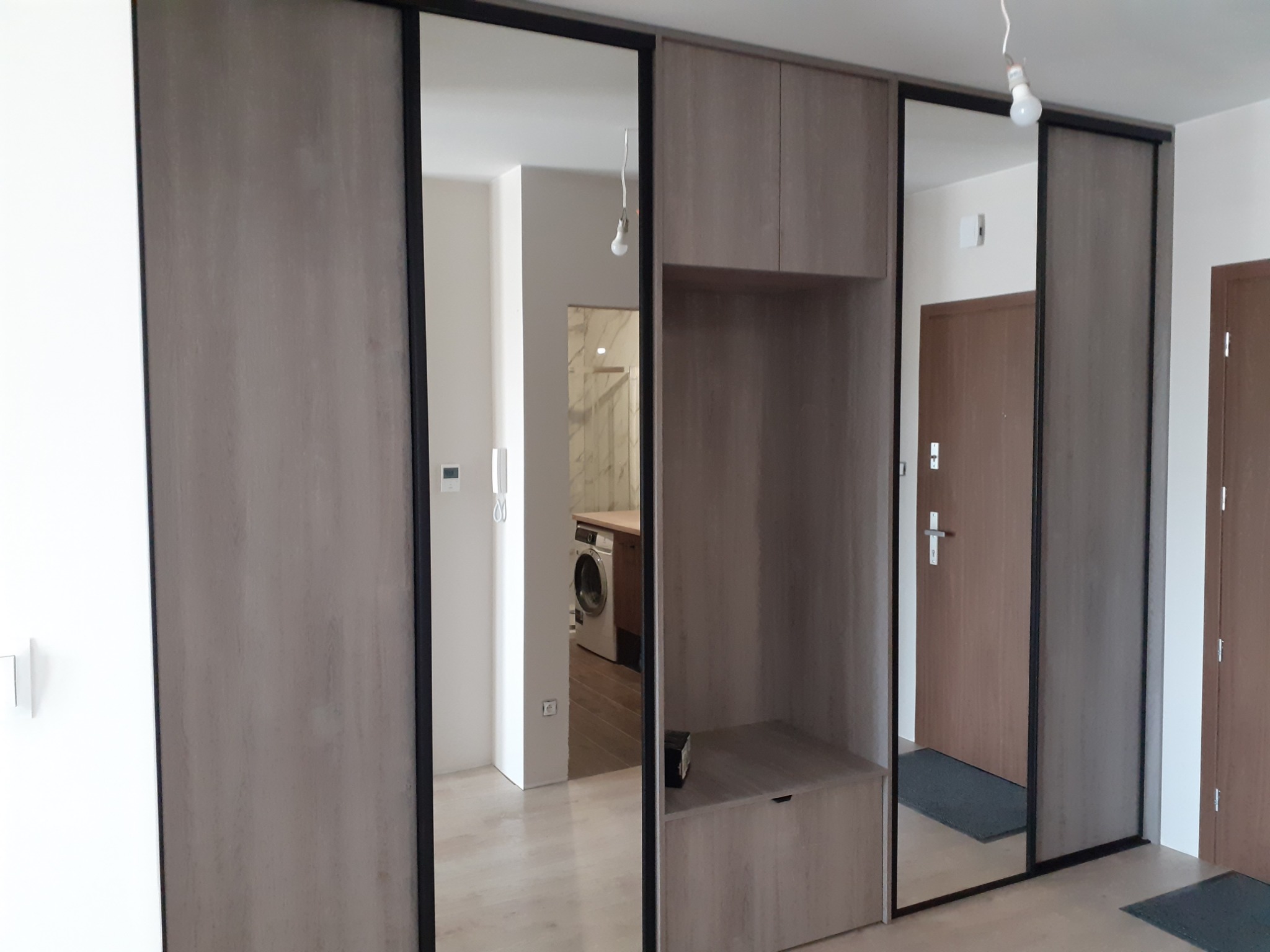 Szafa wnękowa z przesuwnymi drzwiami lustrzanymi i drewnopodobnym wykończeniem, w tle widoczna pralka w łazience.