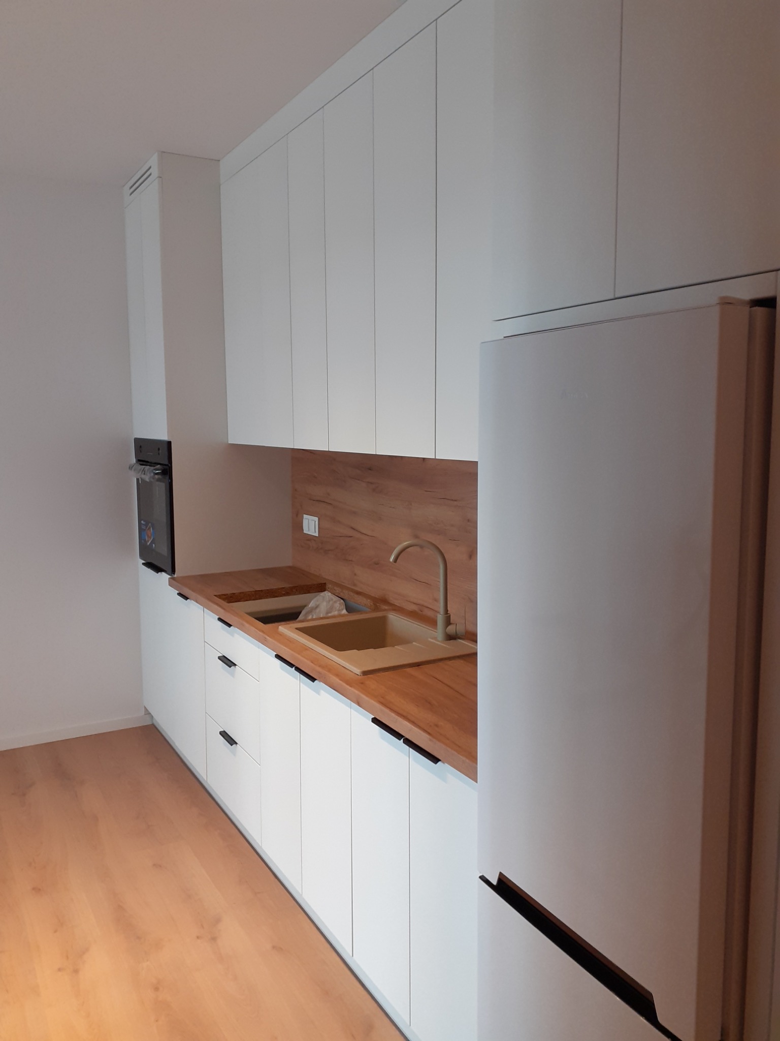 Nowoczesna kuchnia z białymi szafkami z długimi, czarnymi uchwytami, drewnianym blatem i panelem ściennym w odcieniach brązu, oraz beżowym zlewem i baterią, obok stoi biała lodówka.