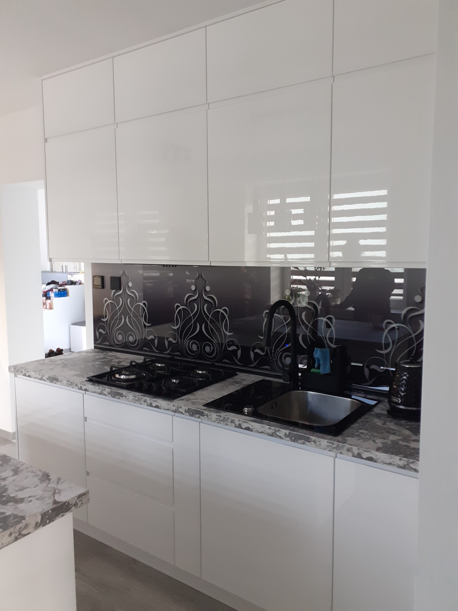 Nowoczesna, biała kuchnia z błyszczącymi frontami szafek górnych i dolnych, szarym blatem imitującym kamień, czarną płytą grzewczą i zlewozmywakiem, oraz dekoracyjnym panelem szklanym nad blatem...