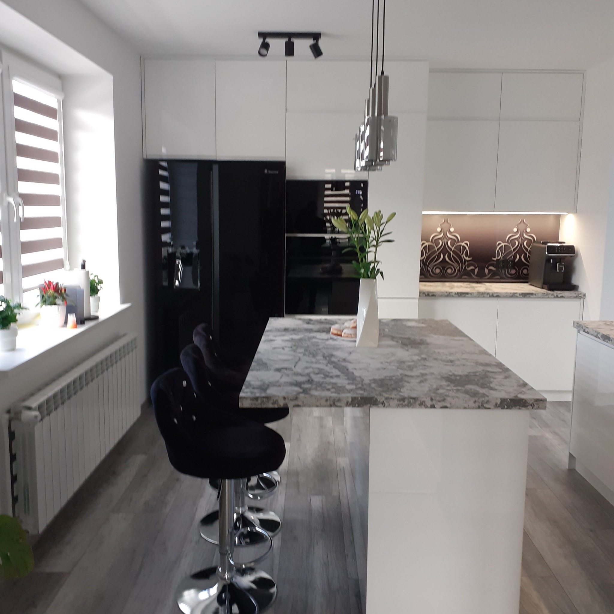 Nowoczesna kuchnia z białymi szafkami, czarną lodówką i wyspą z blatem imitującym marmur, oświetlona trzema lampami punktowymi i wiszącą lampą dekoracyjną.