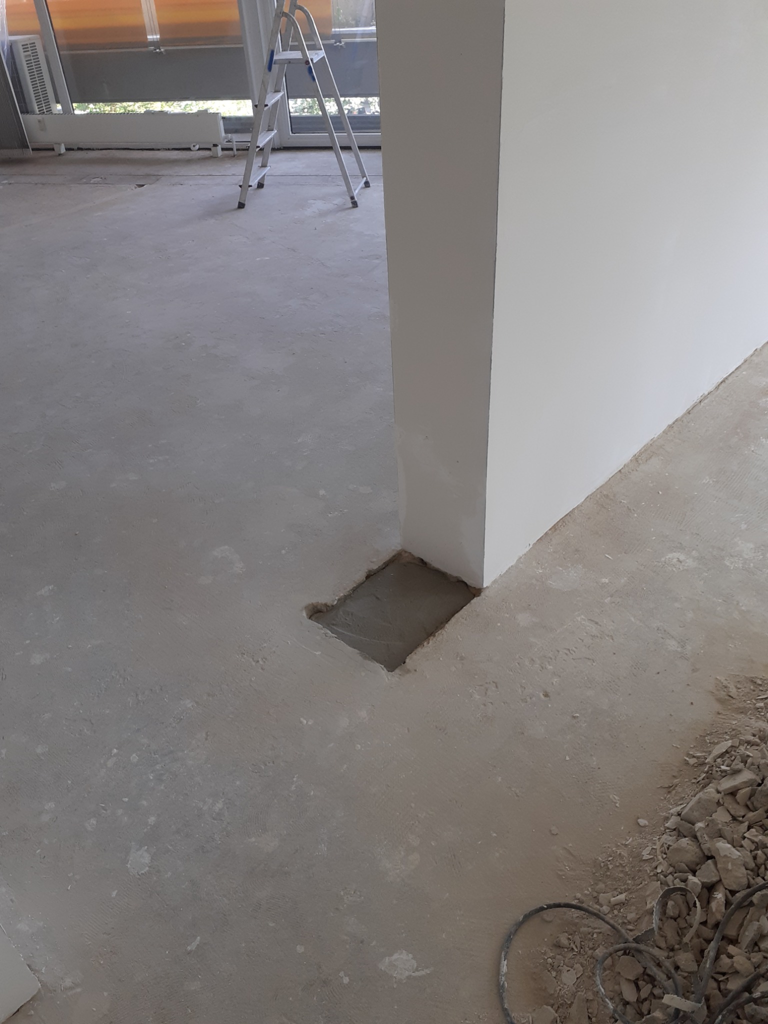 Przygotowanie podłoża: Wyrównana wylewka z wyciętym otworem na instalację przy ścianie, drabina malarska w tle, fragmenty gruzu i kabel na podłodze.