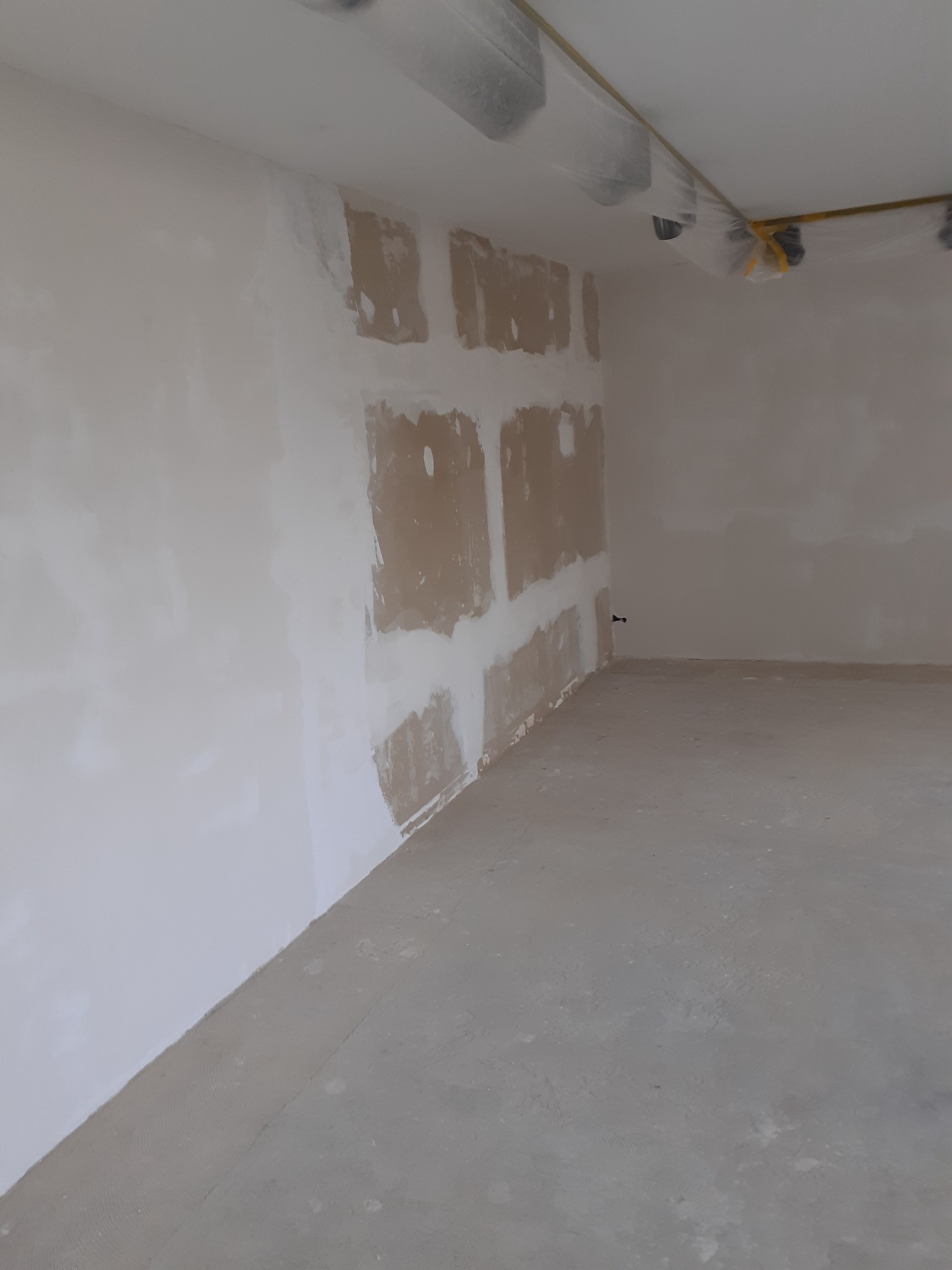 Wnętrze pokoju w trakcie prac wykończeniowych, ściana częściowo pokryta szpachlą w kształcie kwadratów, zabezpieczone lampy na suficie folią ochronną.