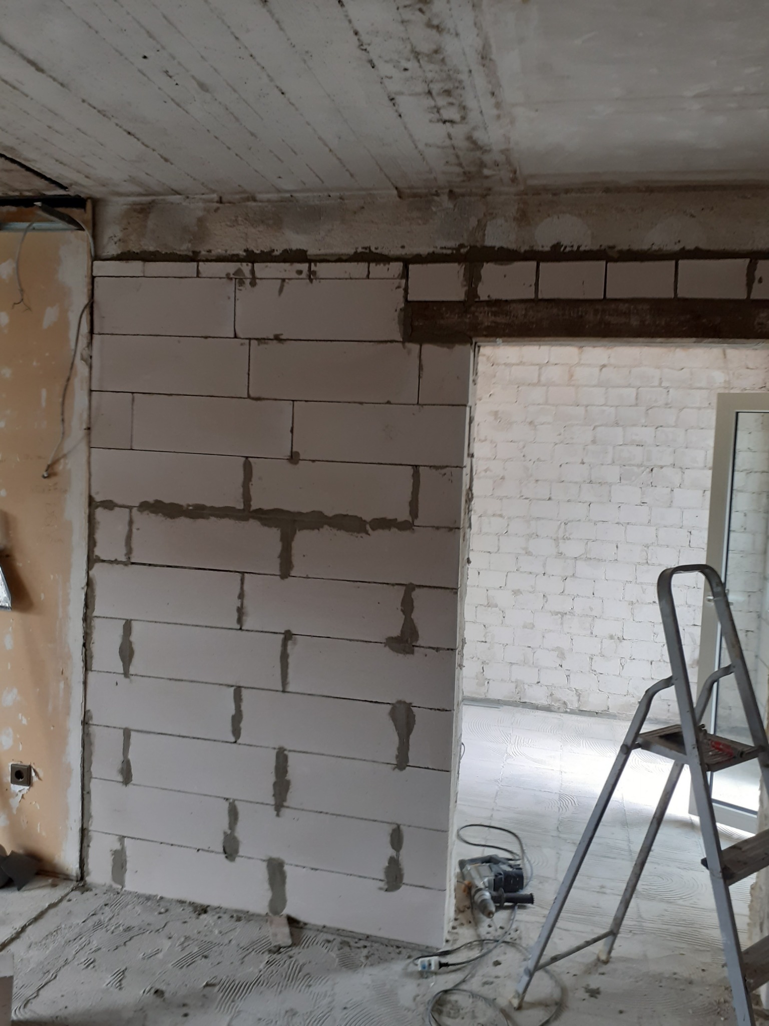 Wnętrze w trakcie rozbudowy: Ściana z białych bloczków, częściowo otynkowana, z widocznym otworem drzwiowym. Na podłodze leży wiertarka udarowa i drabina aluminiowa.