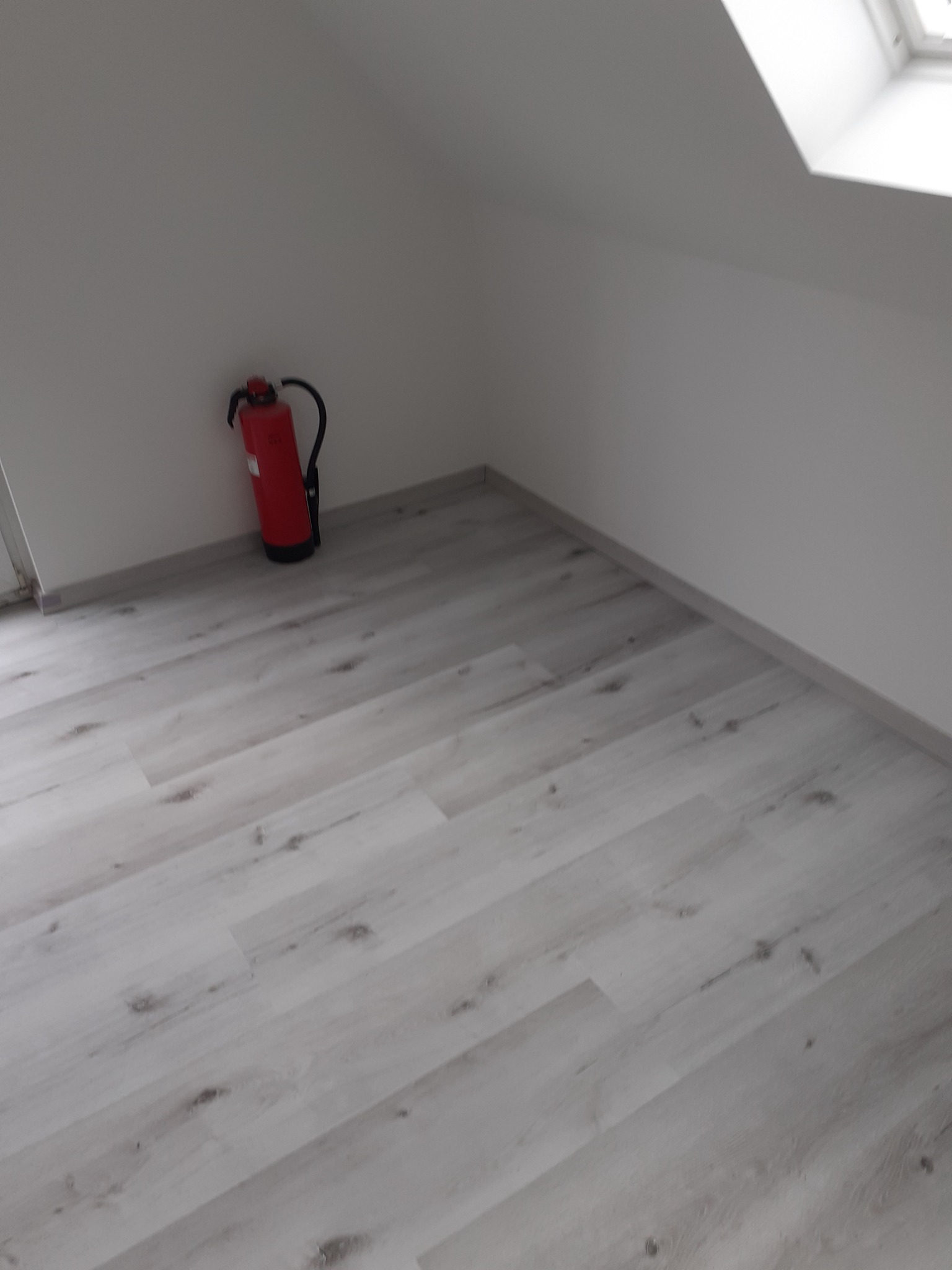 Jasne panele podłogowe w pokoju na poddaszu z oknem dachowym, widoczny czerwony gaśnica przy ścianie.