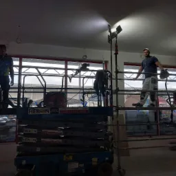 Dw&oacute;ch pracownik&oacute;w na podnośniku i rusztowaniu podczas szpachlowania i wygładzania sufitu w dużym pomieszczeniu, oświetlonych reflektorami.