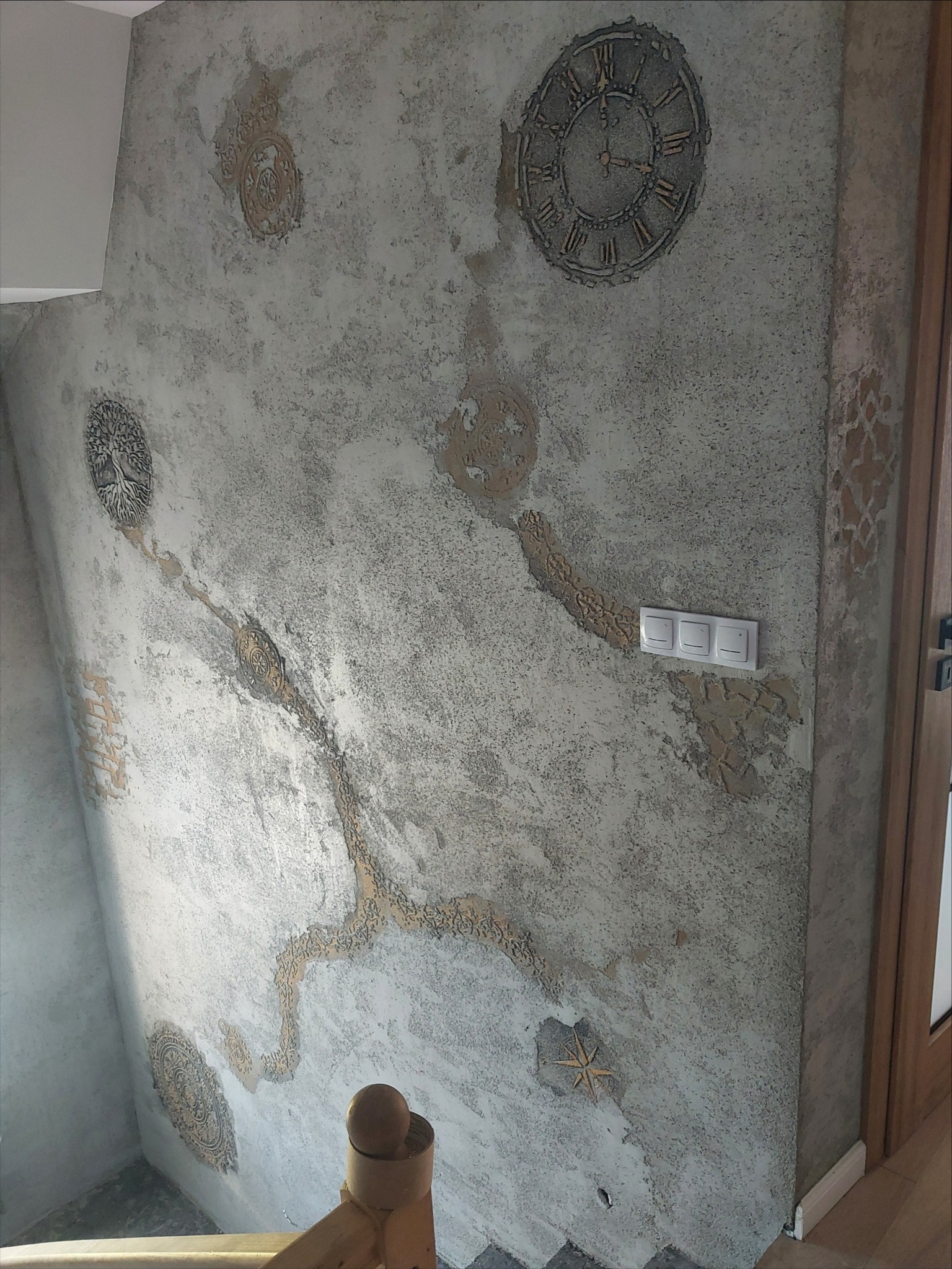 Ściana w klatce schodowej z dekoracyjną, szarą fakturą i wypukłymi, złotymi ornamentami: zegar, drzewo, rozety, gwiazda, wzór przypominający żyły, obok włącznik światła.