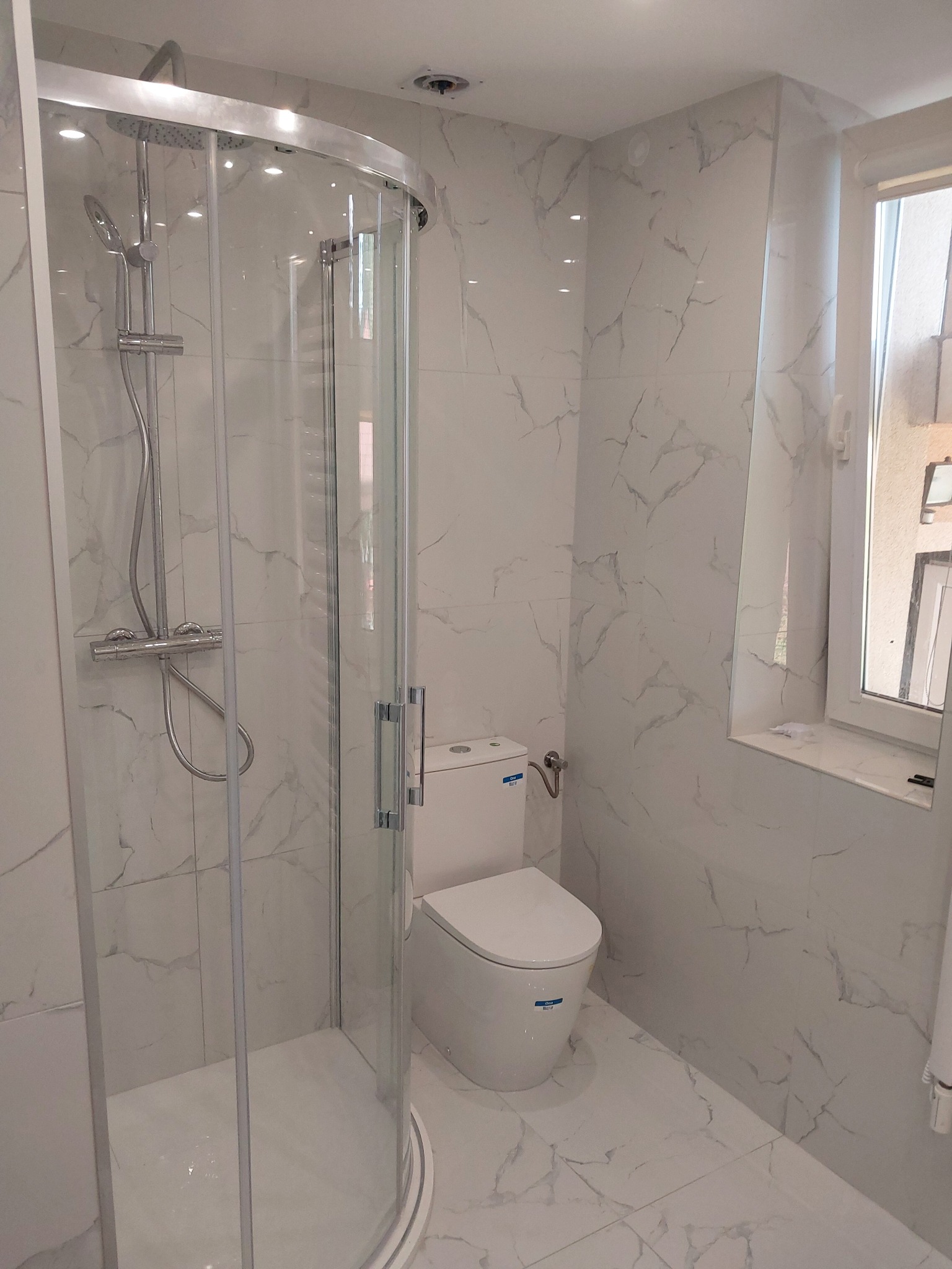 Nowoczesna łazienka z kabiną prysznicową z zaokrąglonym szkłem, toaletą kompaktową i marmurowymi płytkami na ścianach i podłodze.