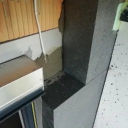 Fragment ściany z widocznym ociepleniem styropianem grafitowym przy oknie, częściowo zamontowanym na klej, z widocznymi elementami siatki zbrojącej przy ościeżnicy.