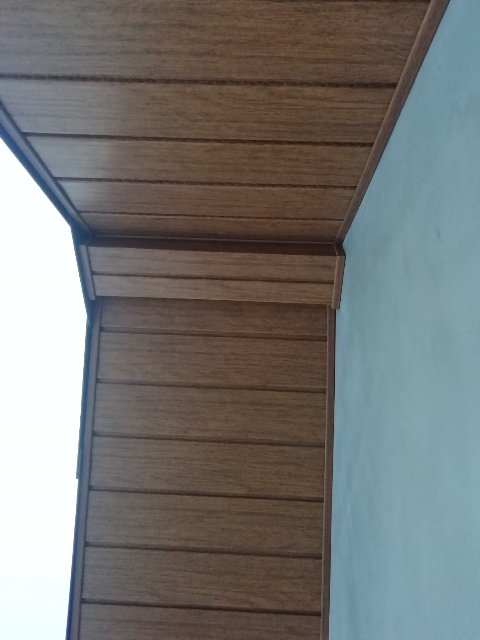 Fragment elewacji z drewnopodobnych paneli, widoczny narożnik budynku i fragment ściany pomalowanej na jasny kolor.