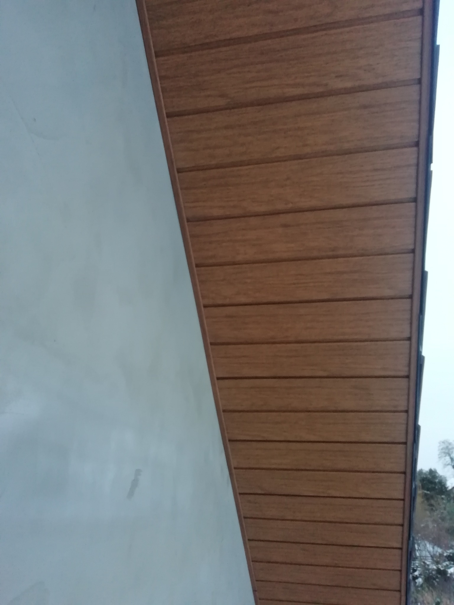 Fragment elewacji: surowa, jasna ściana sąsiaduje z brązową, drewnopodobną podbitką dachową, widok z dołu.