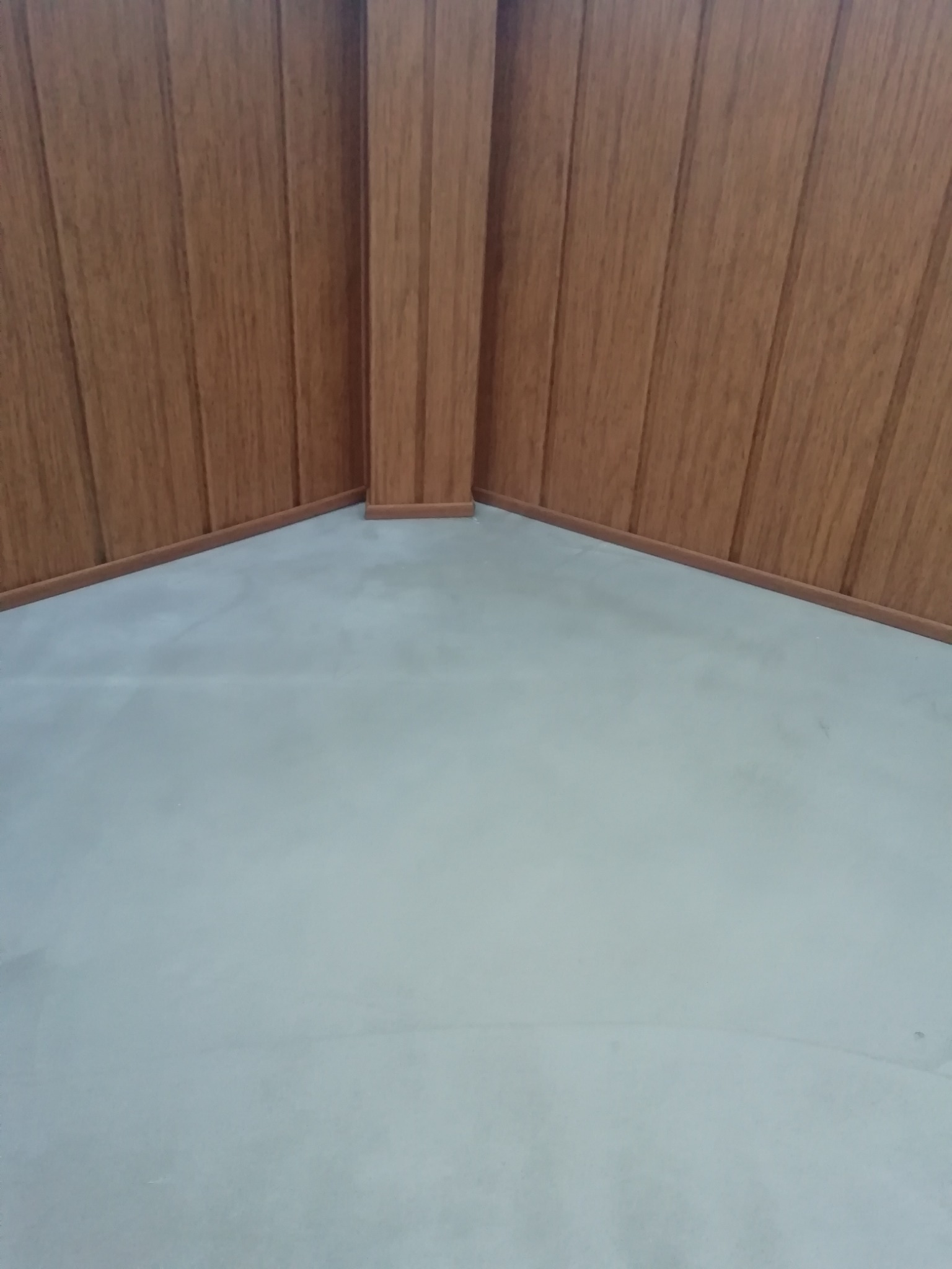 Szary betonowy podkład podłogowy w rogu pomieszczenia z boazerią imitującą drewno na ścianach.