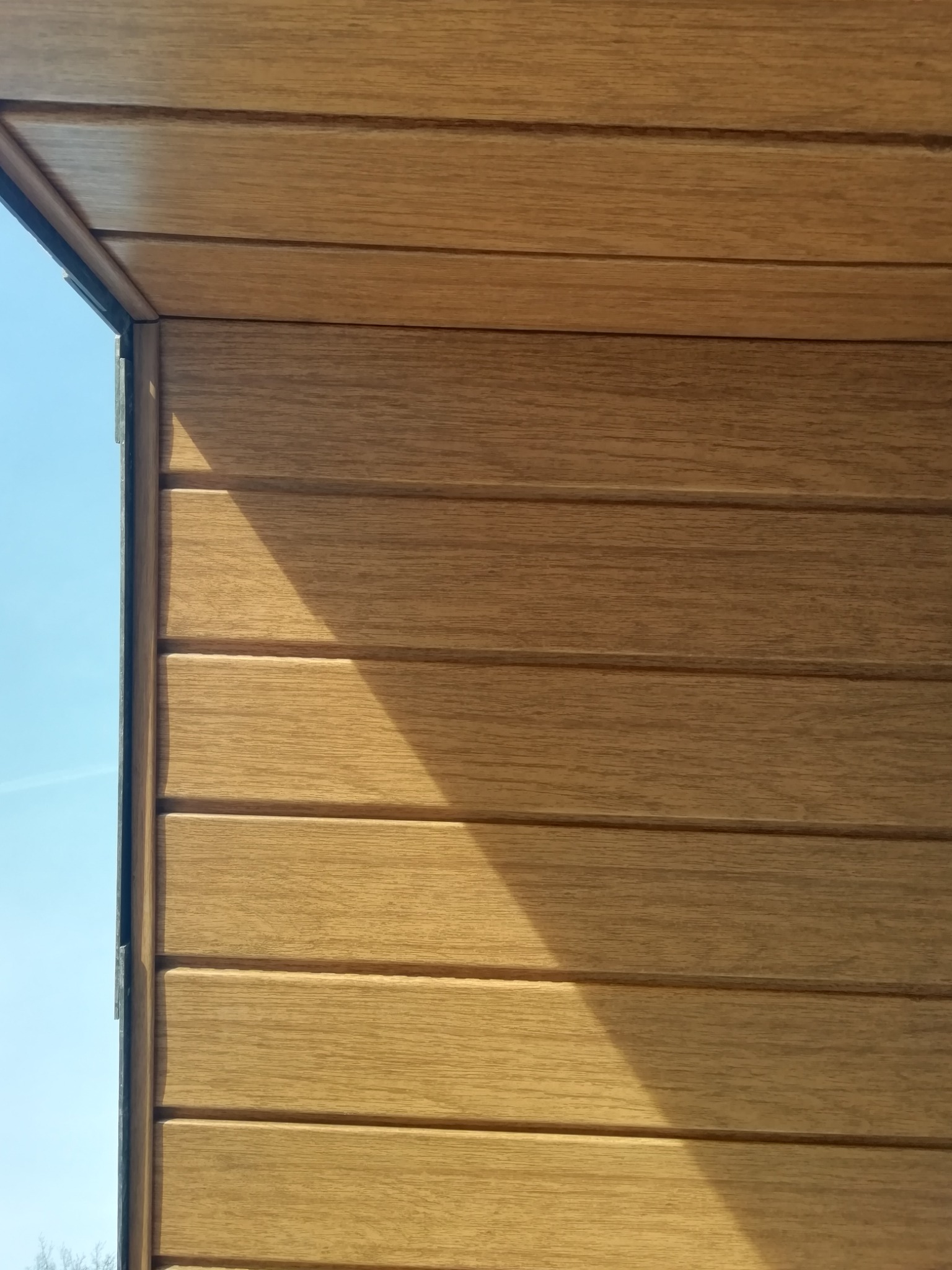 Fragment elewacji z paneli drewnopodobnych, widoczny narożnik budynku i błękitne niebo