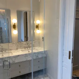 Nowoczesna łazienka z marmurowymi powierzchniami, szklaną kabiną prysznicową z przesuwnymi drzwiami i podwójną umywalką z szafką w kolorze szarym, oświetlona kulistymi lampami.