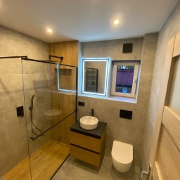 Marek Kimborowicz - Nowoczesna łazienka z kabiną typu walk-in, umywalką nablatową na drewnianej szafce i podświetlanym lustrem. Szare płytki, czarne akcenty i drewniane elementy dekoracyjne.