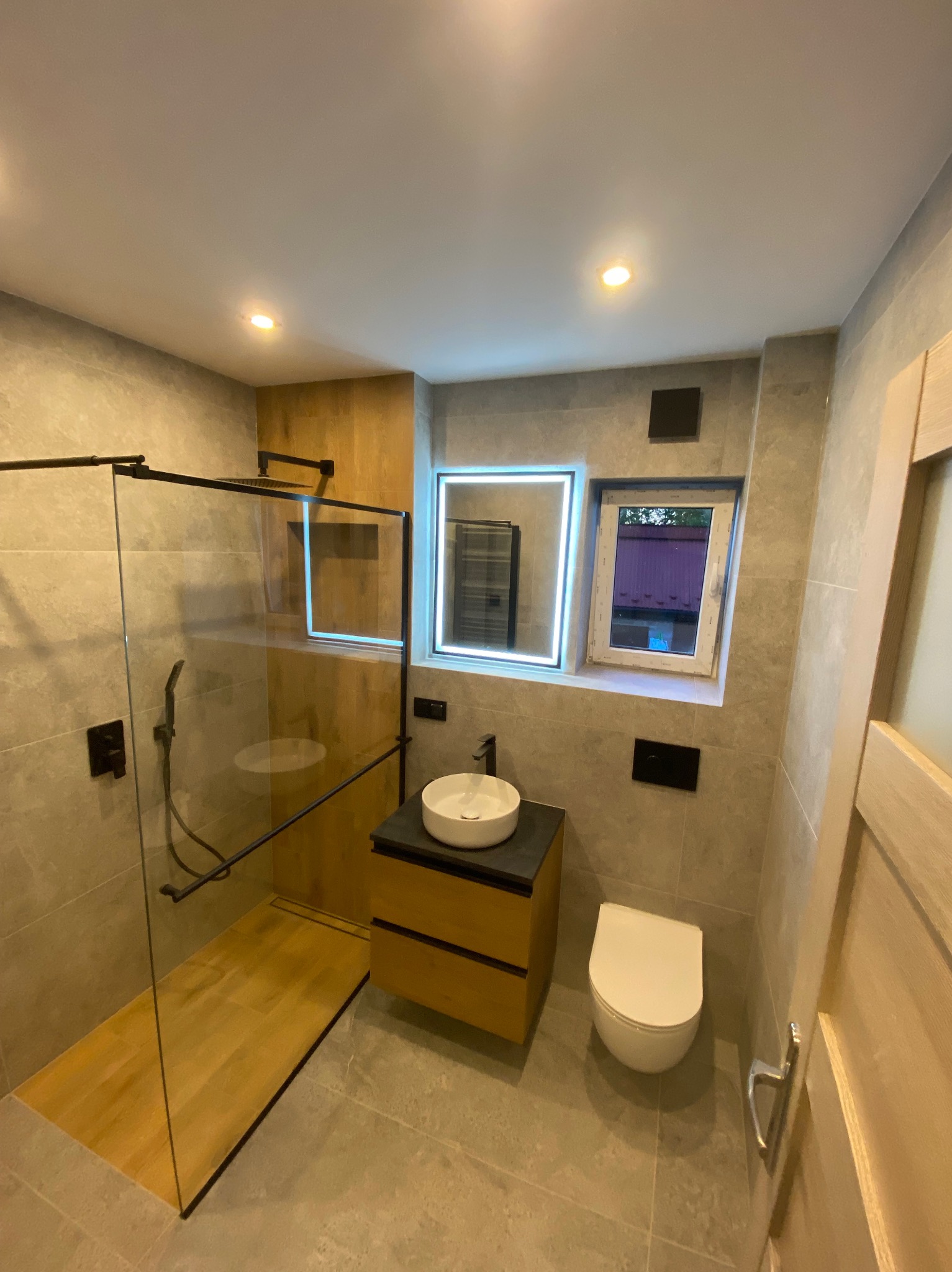 Nowoczesna łazienka z kabiną typu walk-in, umywalką nablatową na drewnianej szafce i podświetlanym lustrem. Szare płytki, czarne akcenty i drewniane elementy dekoracyjne.