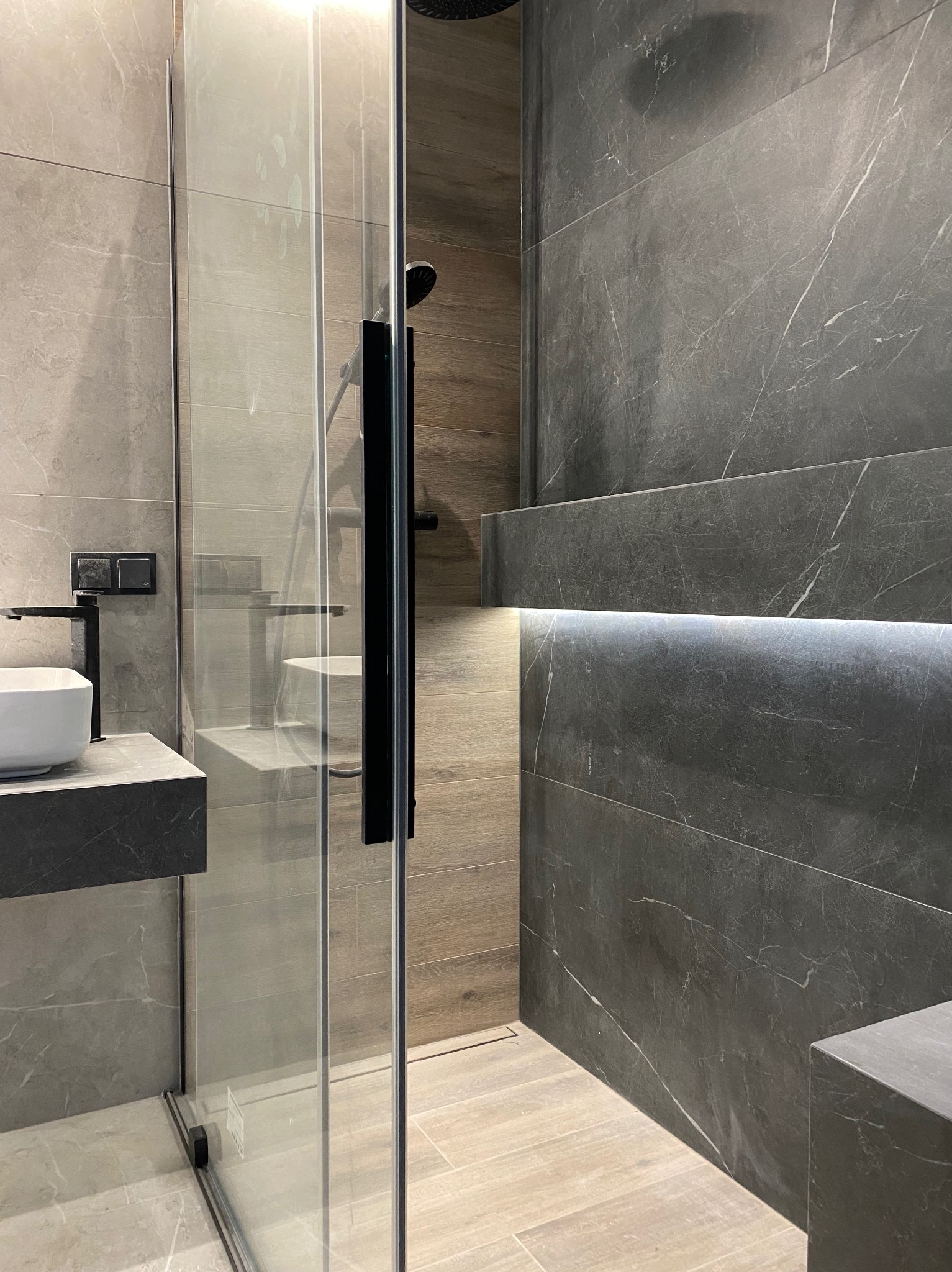 Nowoczesna łazienka z szarymi płytkami imitującymi marmur, kabiną prysznicową ze szkła i czarnymi detalami oraz podświetlaną półką.